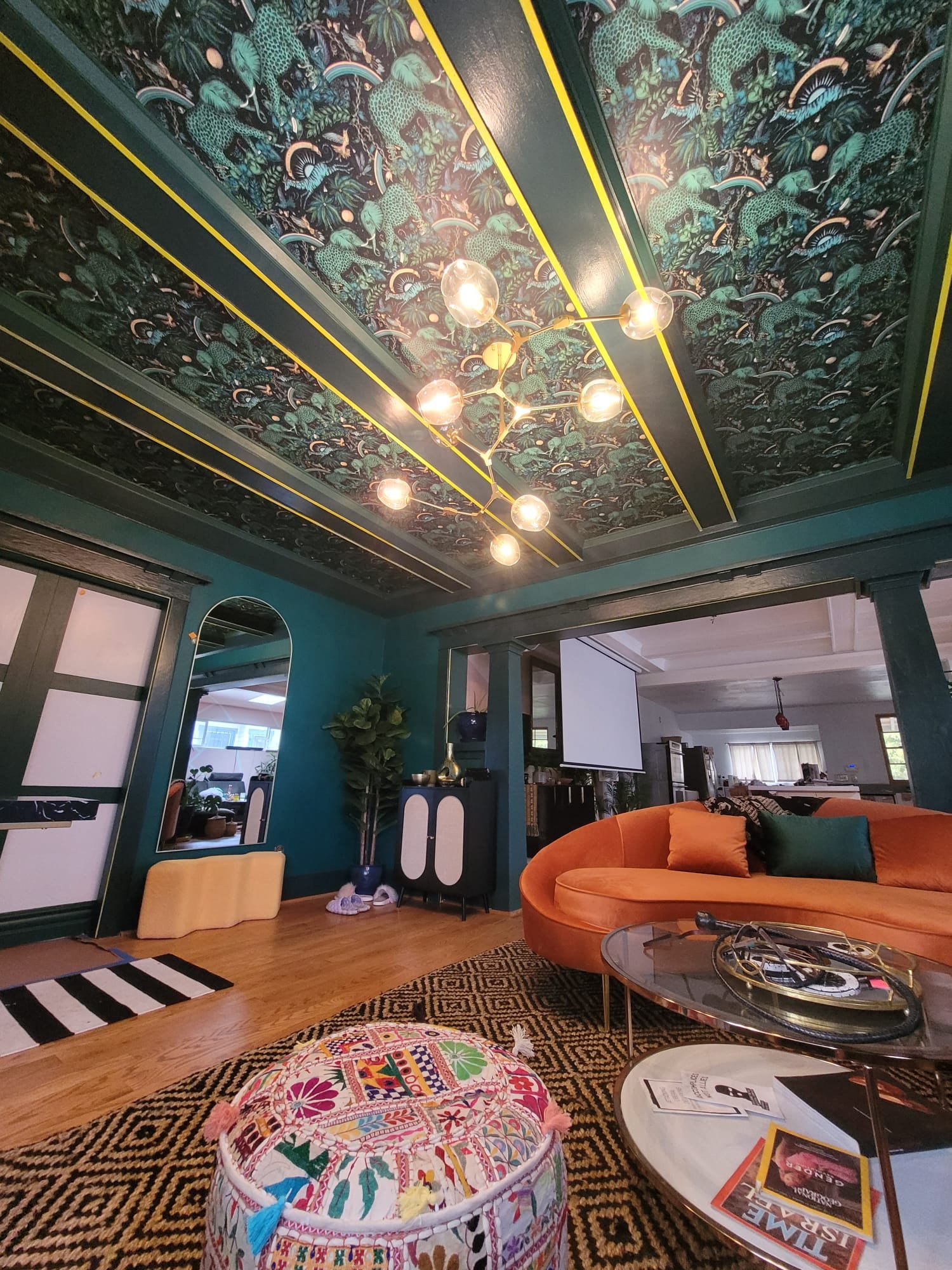 A cozy living room with a colorful and patterned ceiling that features leopards, rainbows, and lush foliage. There's a stylish gold chandelier hanging from the ceiling. The room has blue walls, a teal cabinet, a large orange velvet sectional sofa wit