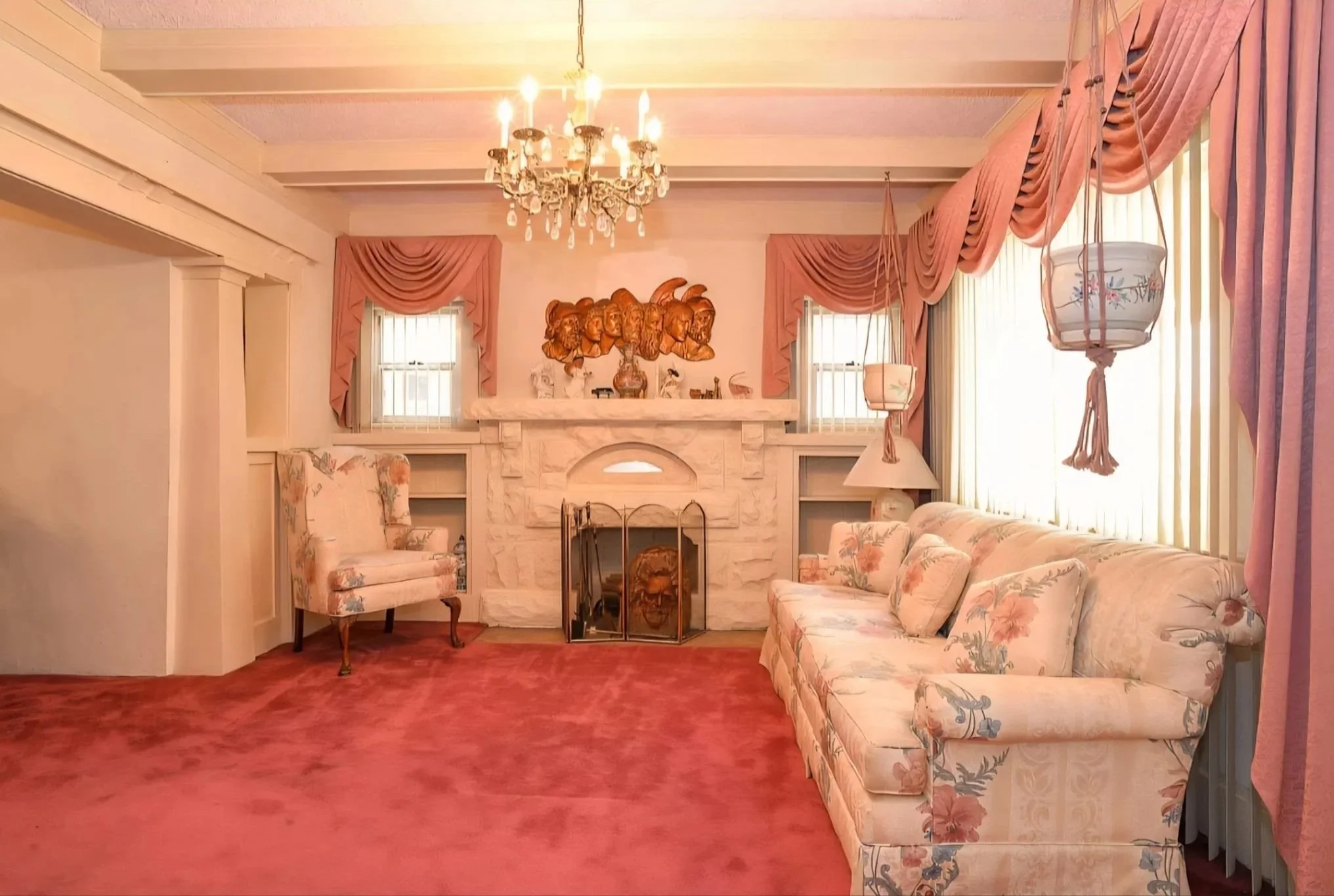A living room with pink floral-patterned upholstery on a sofa and chair, pink curtains, a chandelier, a fireplace with decorations on the mantle, and artwork on the wall above the fireplace.