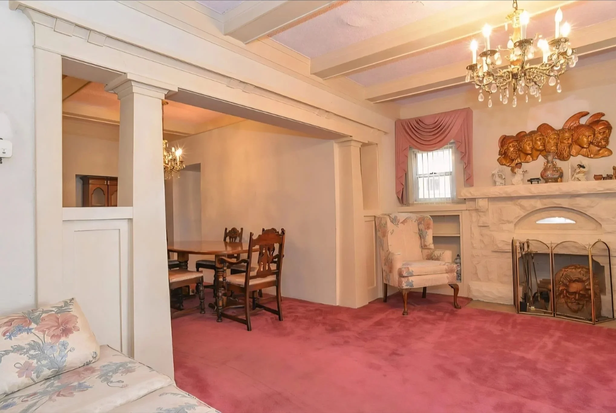 Interior of a living room with pink carpet, cream-colored walls, a marble fireplace, and traditional furniture including floral armchairs, a wooden dining table with chairs, and a chandelier hanging from the ceiling.