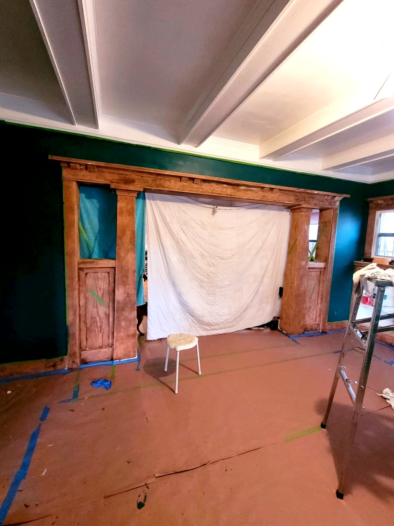 A room under renovation with a wooden doorway framing a space covered by a white sheet. There is a small white stool in front of the doorway, a ladder on the right, and blue painter's tape on the floor.