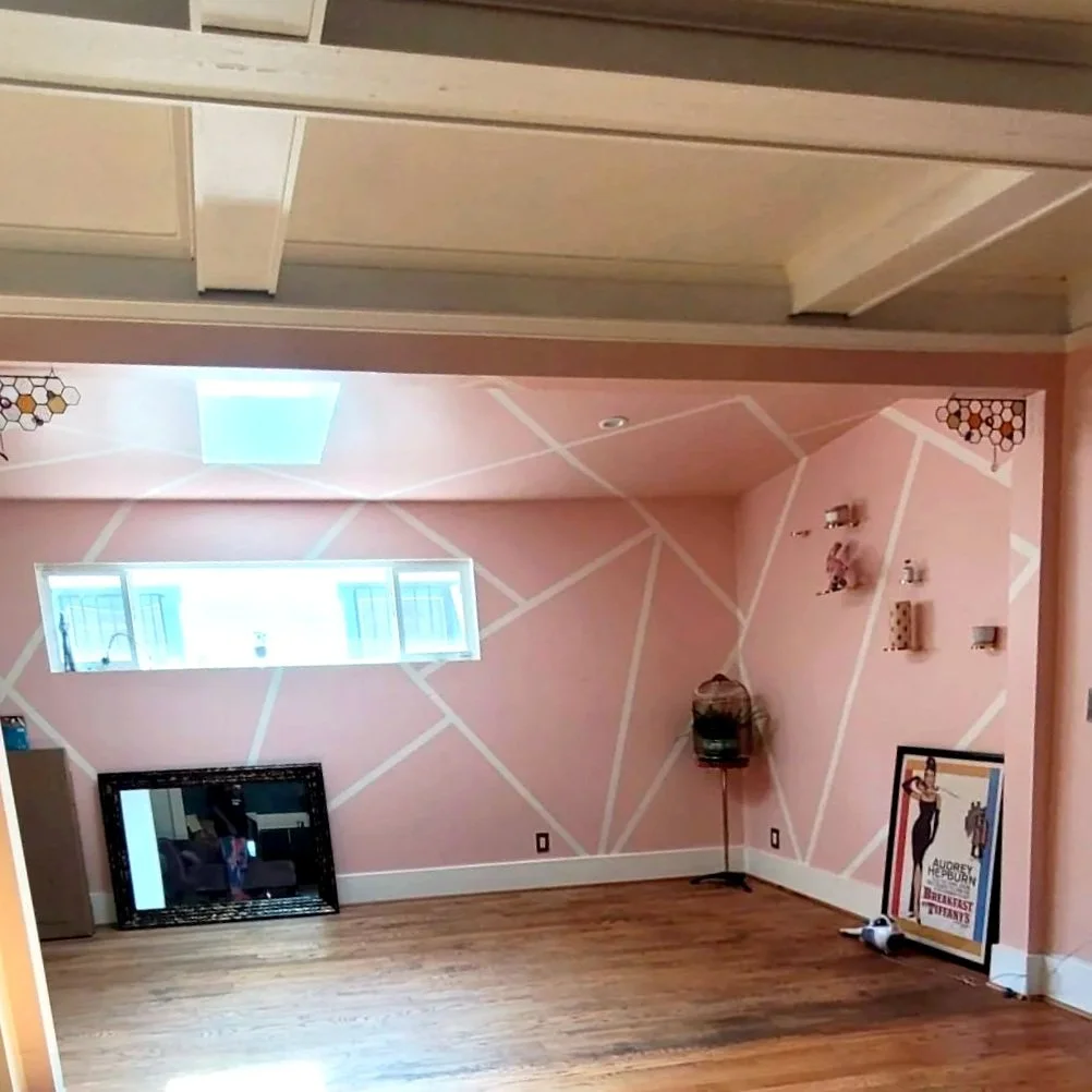 Empty room with pink geometric-patterned wall, a narrow horizontal window, a large mirror leaning against the wall, framed poster on the floor, and a basket hanging from a stand.