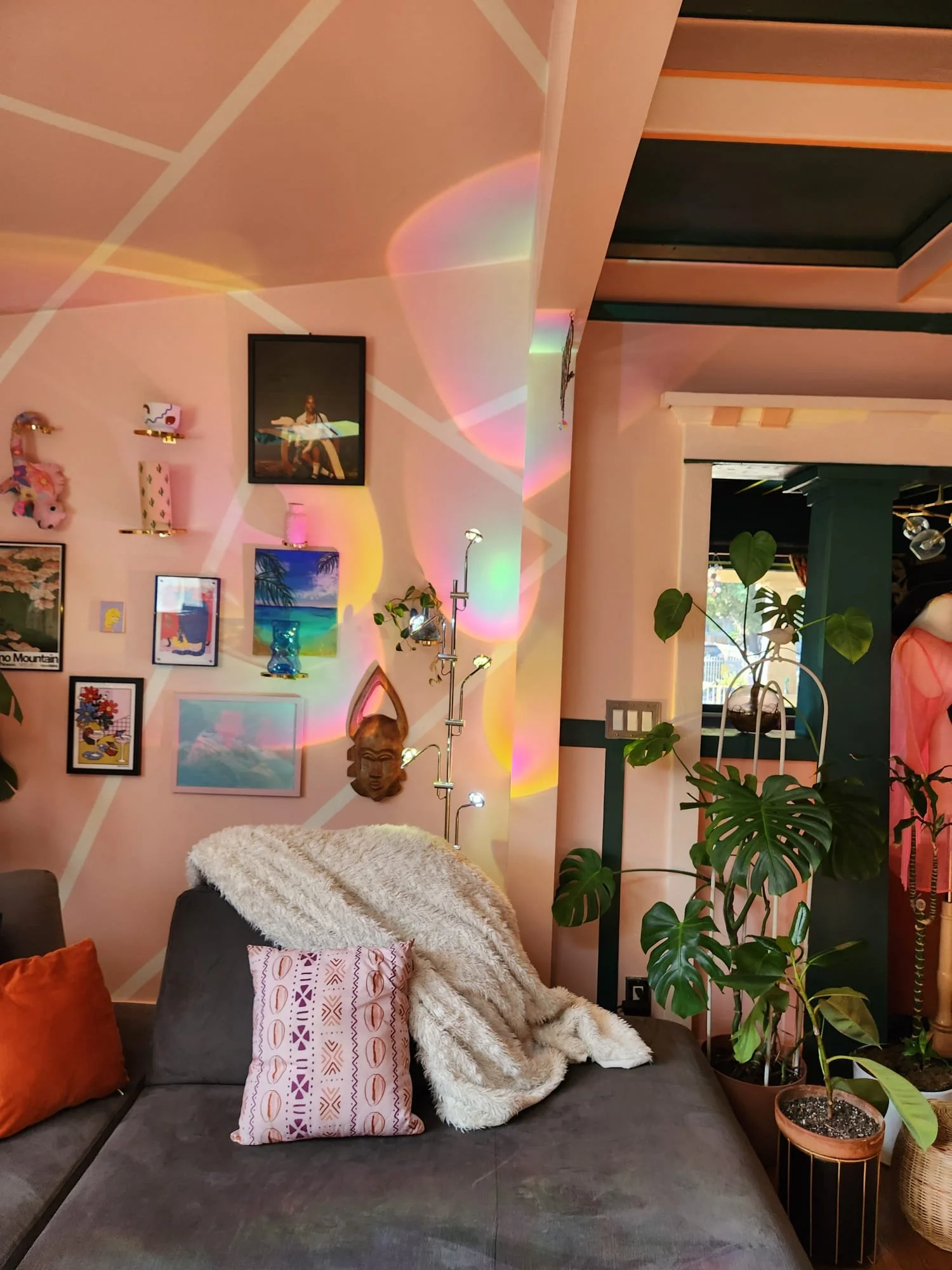 A cozy living room corner with a dark gray sofa, decorative pillows, a plush blanket, and a wall decorated with framed artwork, a mirror, and a wall hanging. There are green plants in pots and colorful light reflections on the wall.