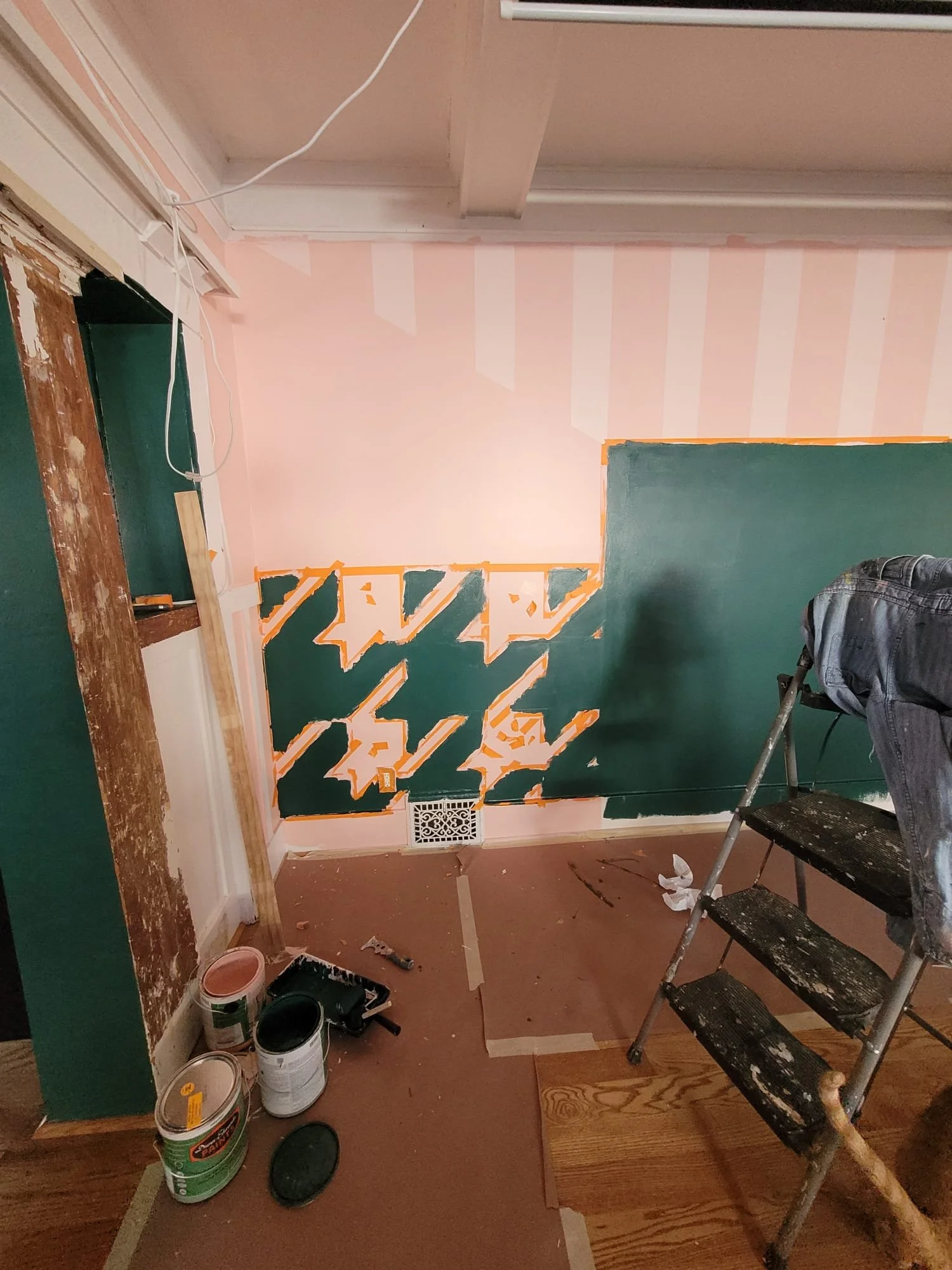 Interior room under renovation with partially painted green wall, pink striped wallpaper, painting supplies on the floor, and a step ladder.