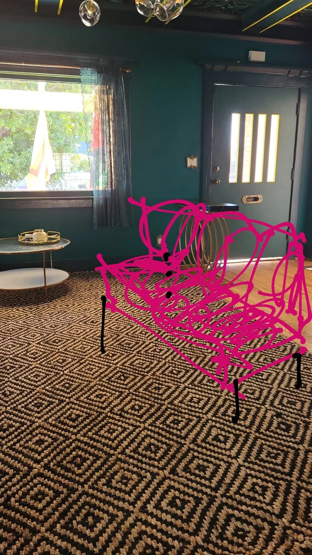 Living room with teal walls, a window with sheer curtains, a patterned rug, a small round table with a tray, and a door with glass panels; pink scribble drawing of a couch overlays the scene.