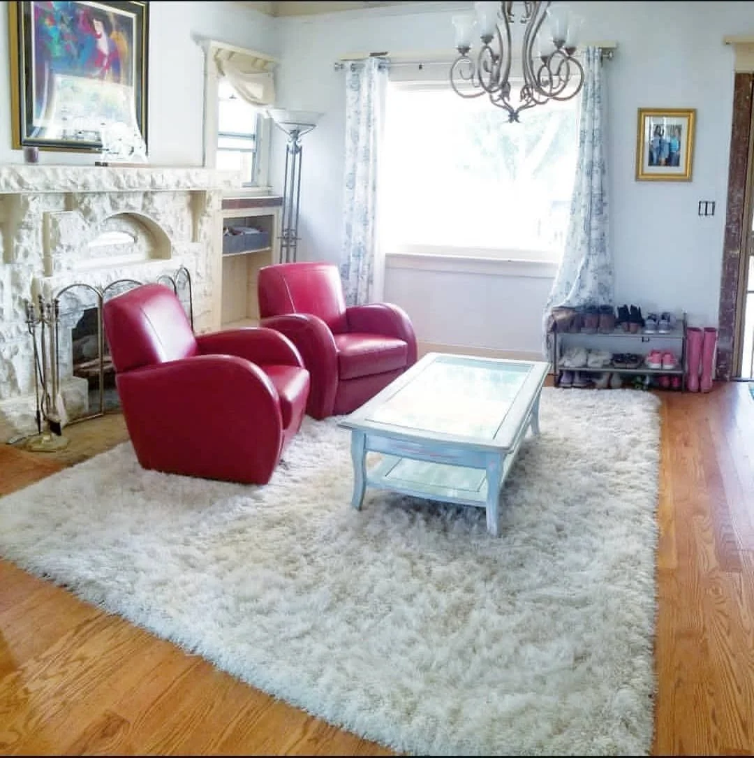 A living room with a white shag rug, two red armchairs, a white coffee table, a stone fireplace, a window with curtains, and a chandelier hanging from the ceiling.