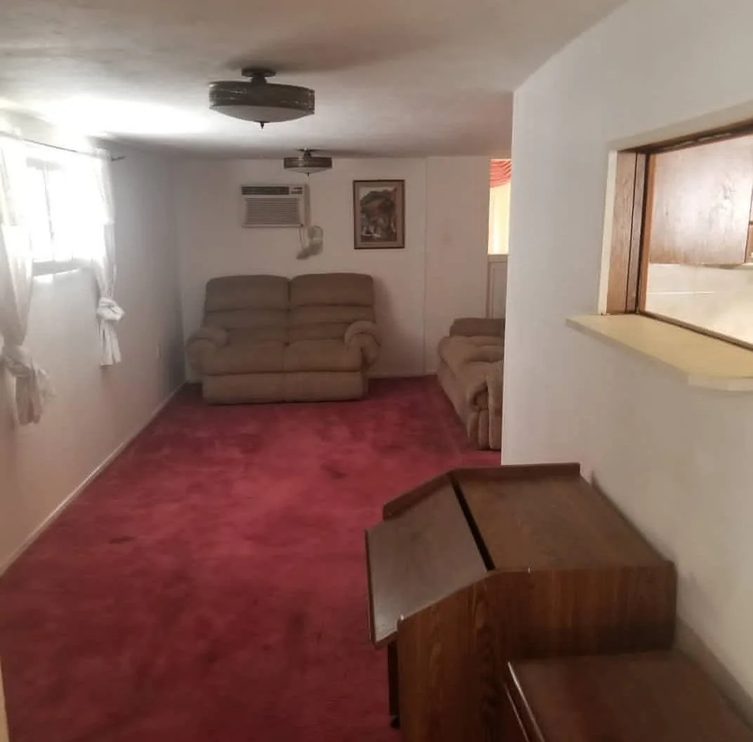 Living room with beige sofas, a window with white curtains, a painting on the wall, a red carpet, and a wooden piano.