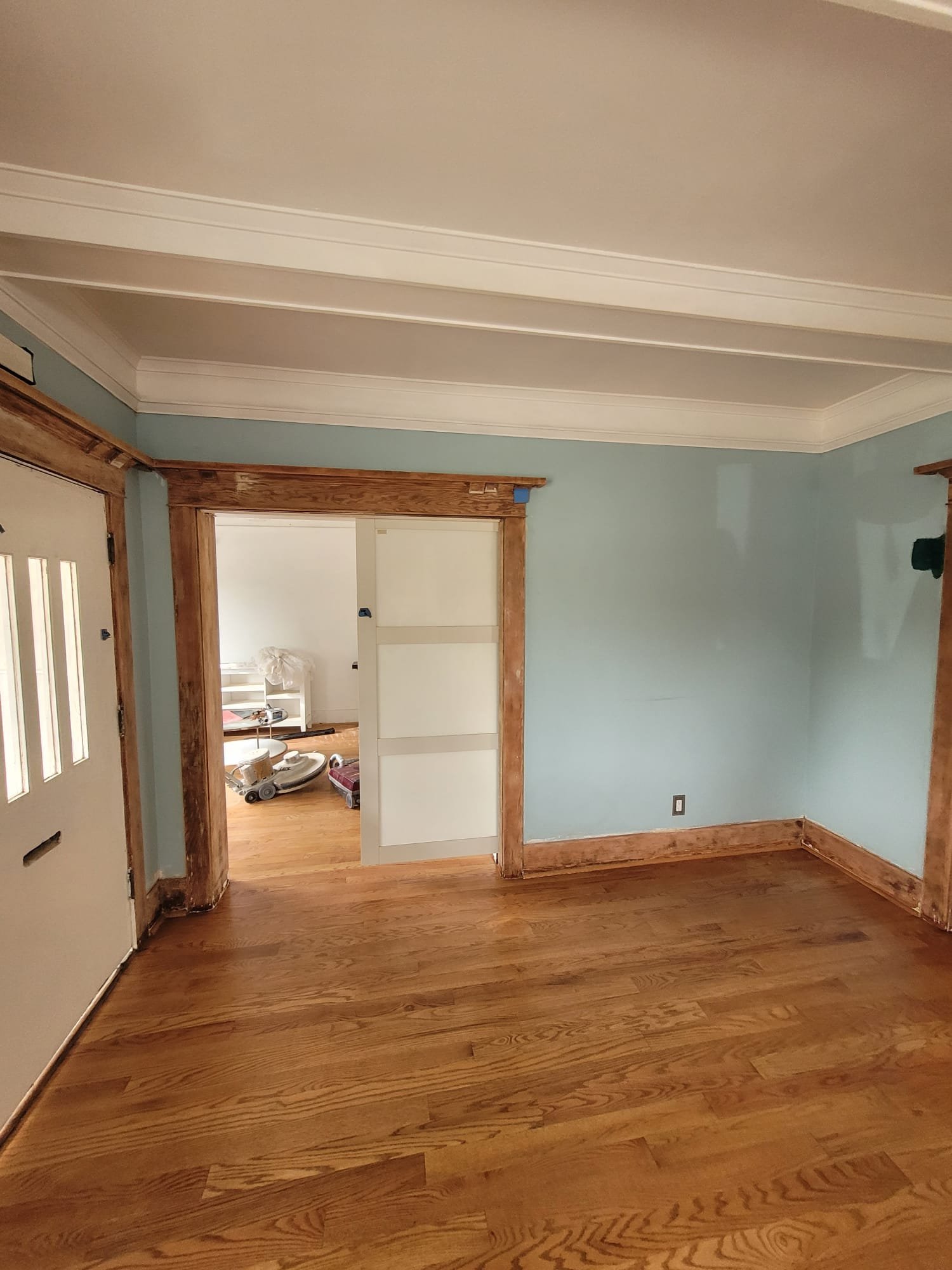 A room with hardwood floor, blue walls, white ceiling with crown molding, wooden door frames, and an open doorway leading to another room with a white wall, some furniture, and tools on the floor.