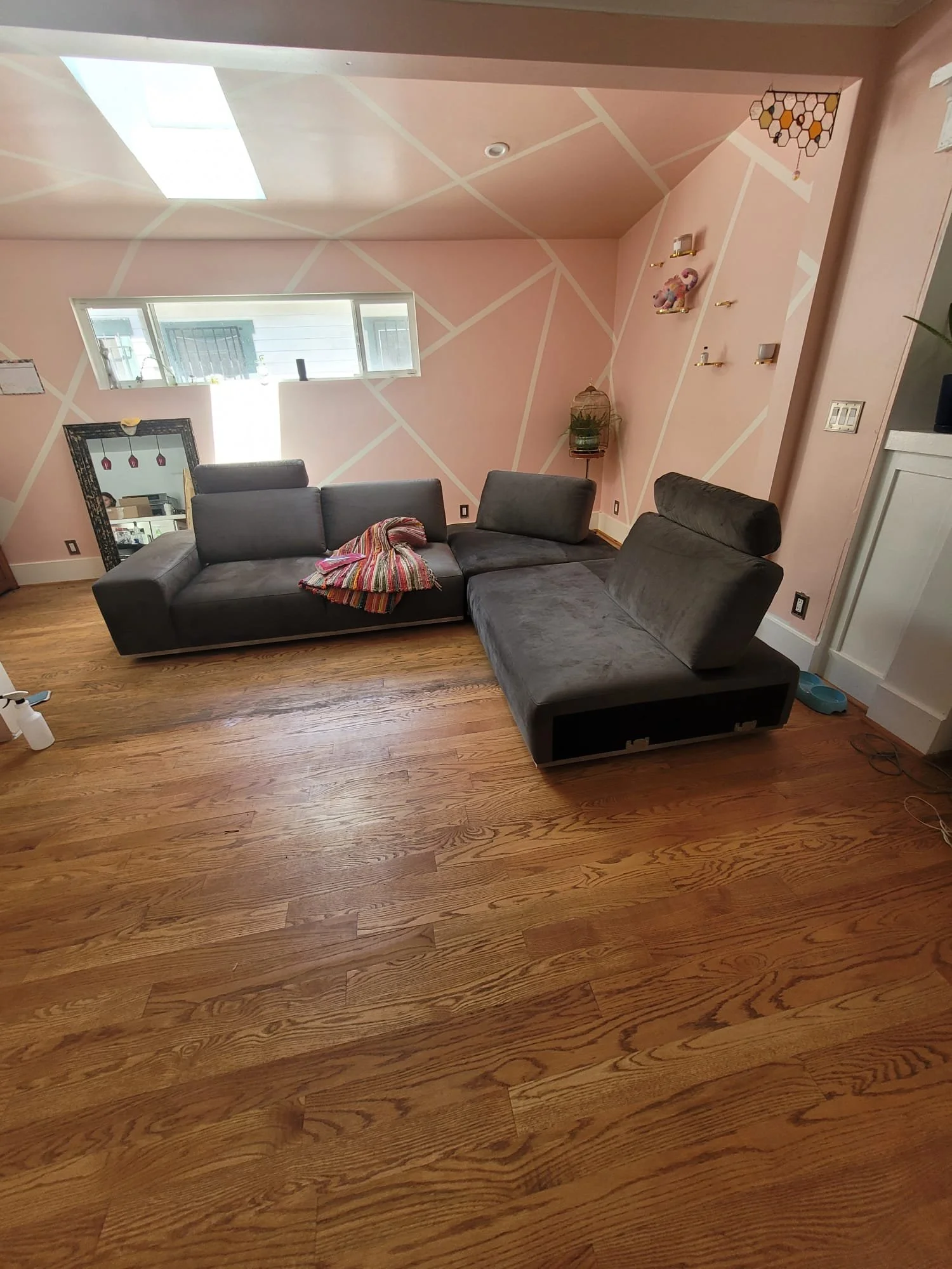 A living room with pink walls decorated with white geometric lines. There is a large window, a black sectional sofa with a multi-colored blanket, a tall mirror, a lamp, and some small decorative shelves. The wooden floor is visible.