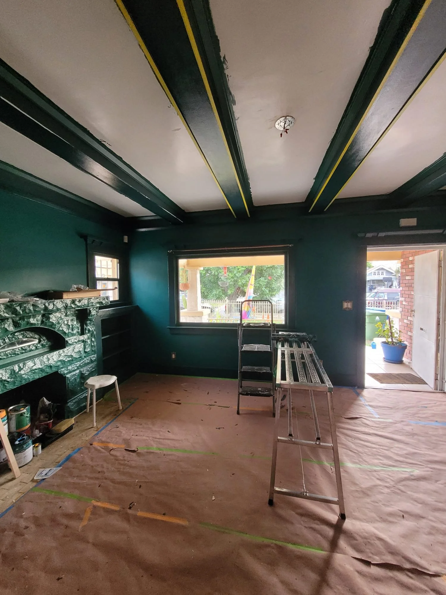 Interior of a room under renovation with dark green walls, a ceiling with exposed beams, and a large window looking out to a backyard. Tools and construction materials are scattered on the floor and furniture.