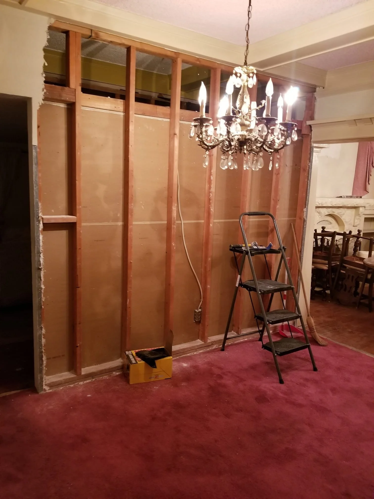 Room with partially removed wall showing wooden studs, chandelier hanging from ceiling, red carpeted floor, ladder, and renovation tools.