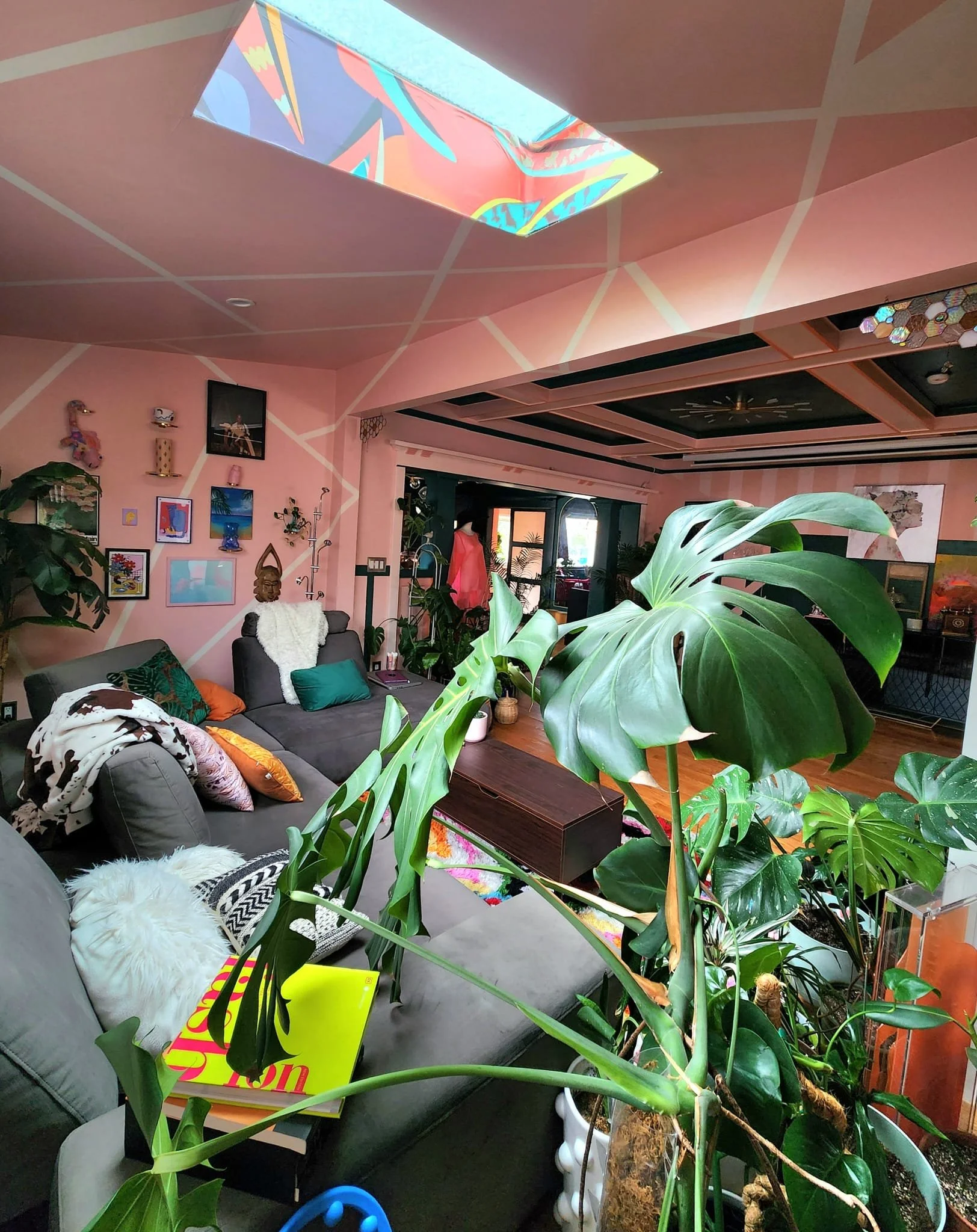A living room with pink walls decorated with art and sculptures, a black sofa with colorful pillows and a white furry throw, large green houseplants in white pots, a wooden coffee table, a wall-mounted TV, and a ceiling with geometric patterns and a 