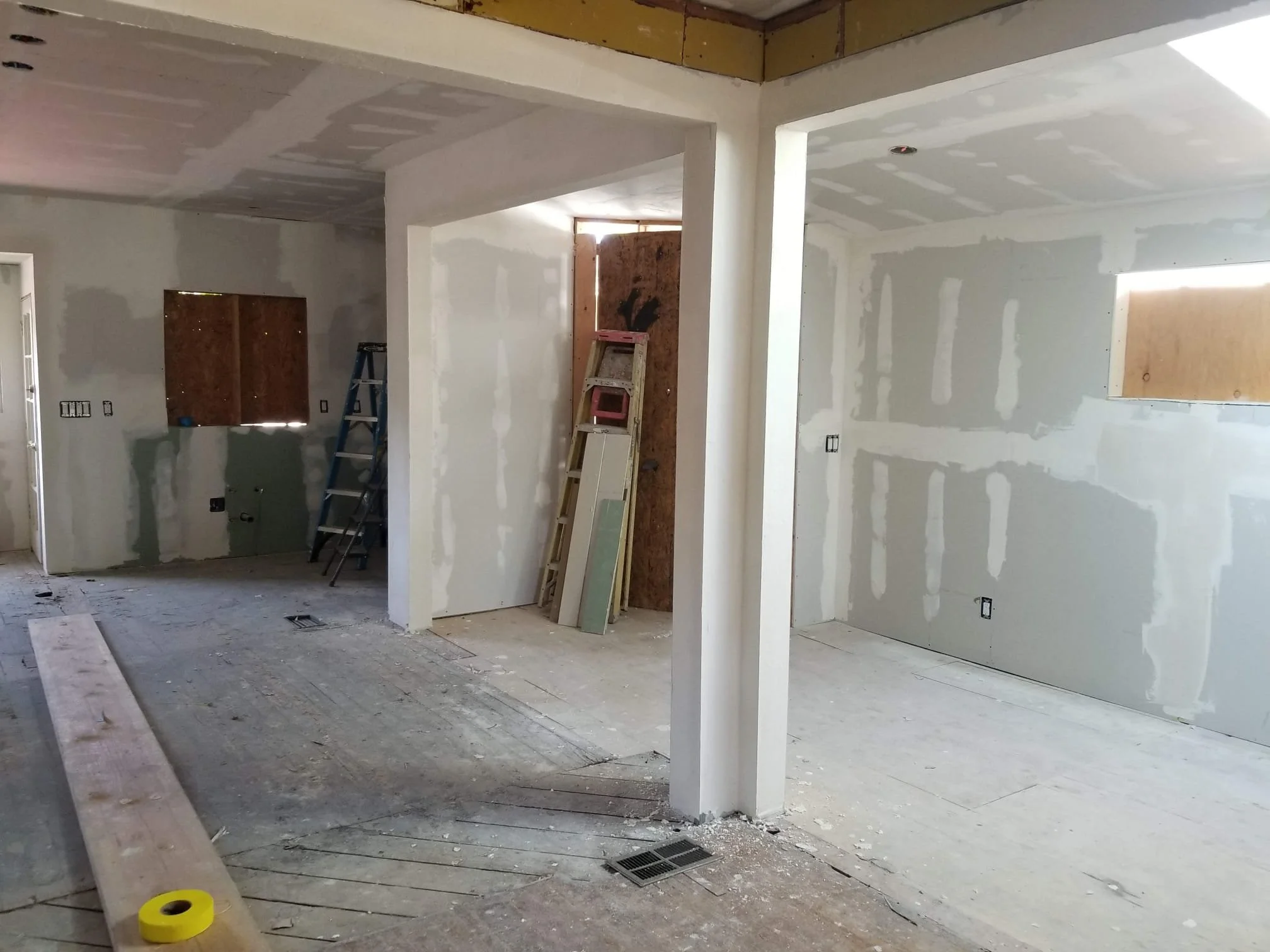 Interior of a house under construction with drywall installation in progress, ladders, and construction tools visible.