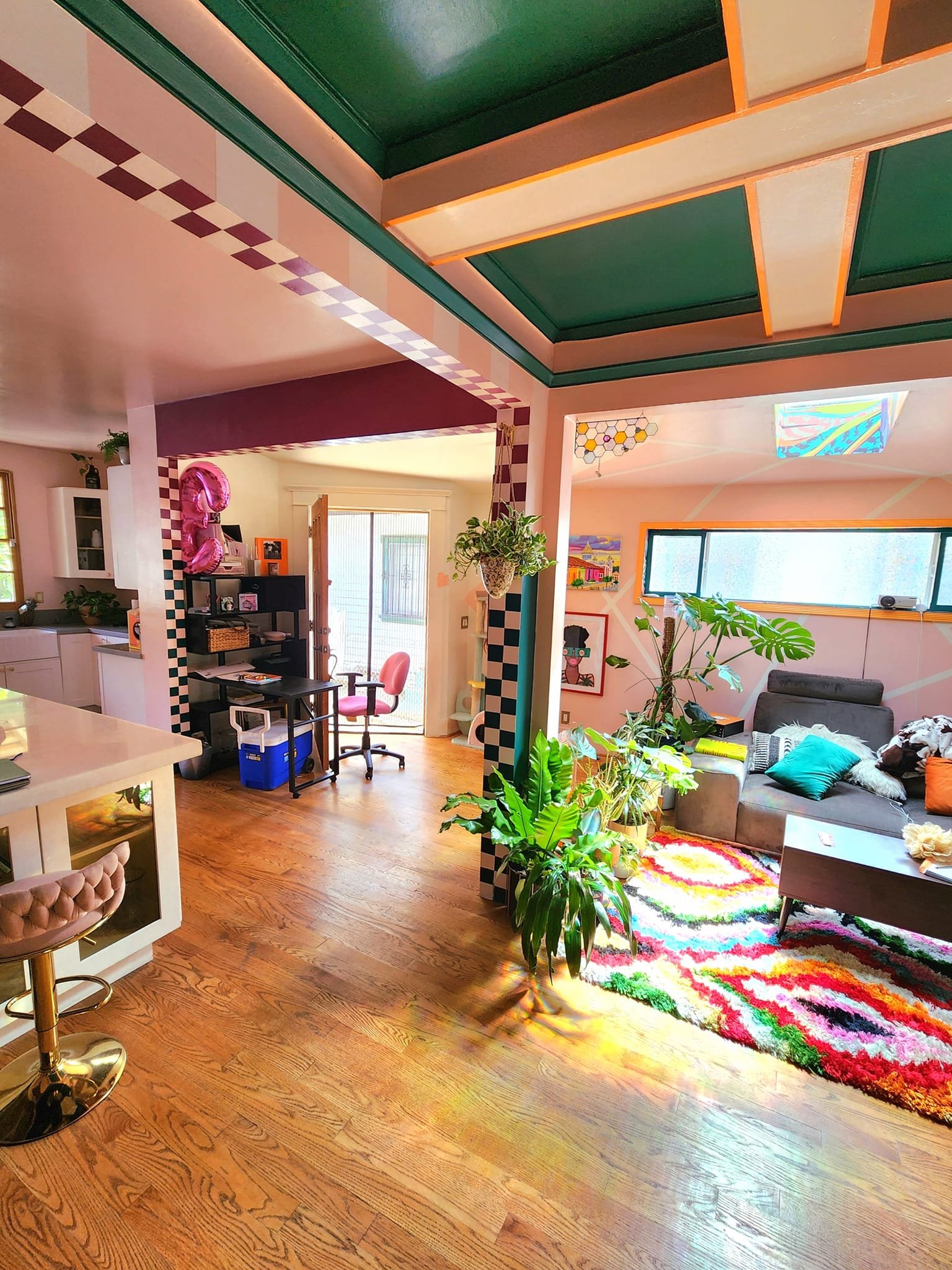 A colorful living room with hardwood floors, a multicolored rug, green plants, a gray sofa, artwork on the walls, and a large window letting in sunlight.
