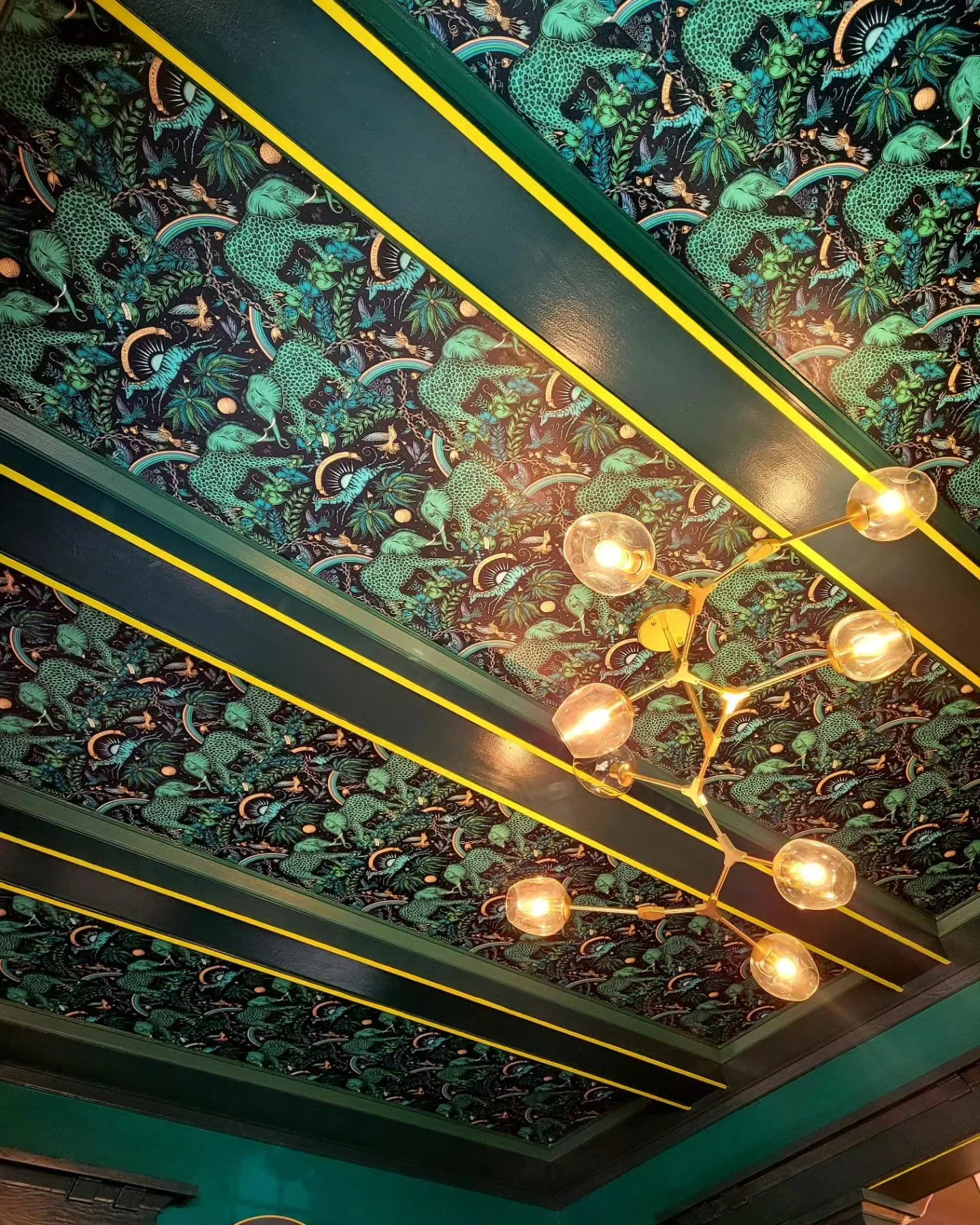 Decorative ceiling with tropical animal and plant pattern, yellow linear accents, and modern chandelier with glass globes.