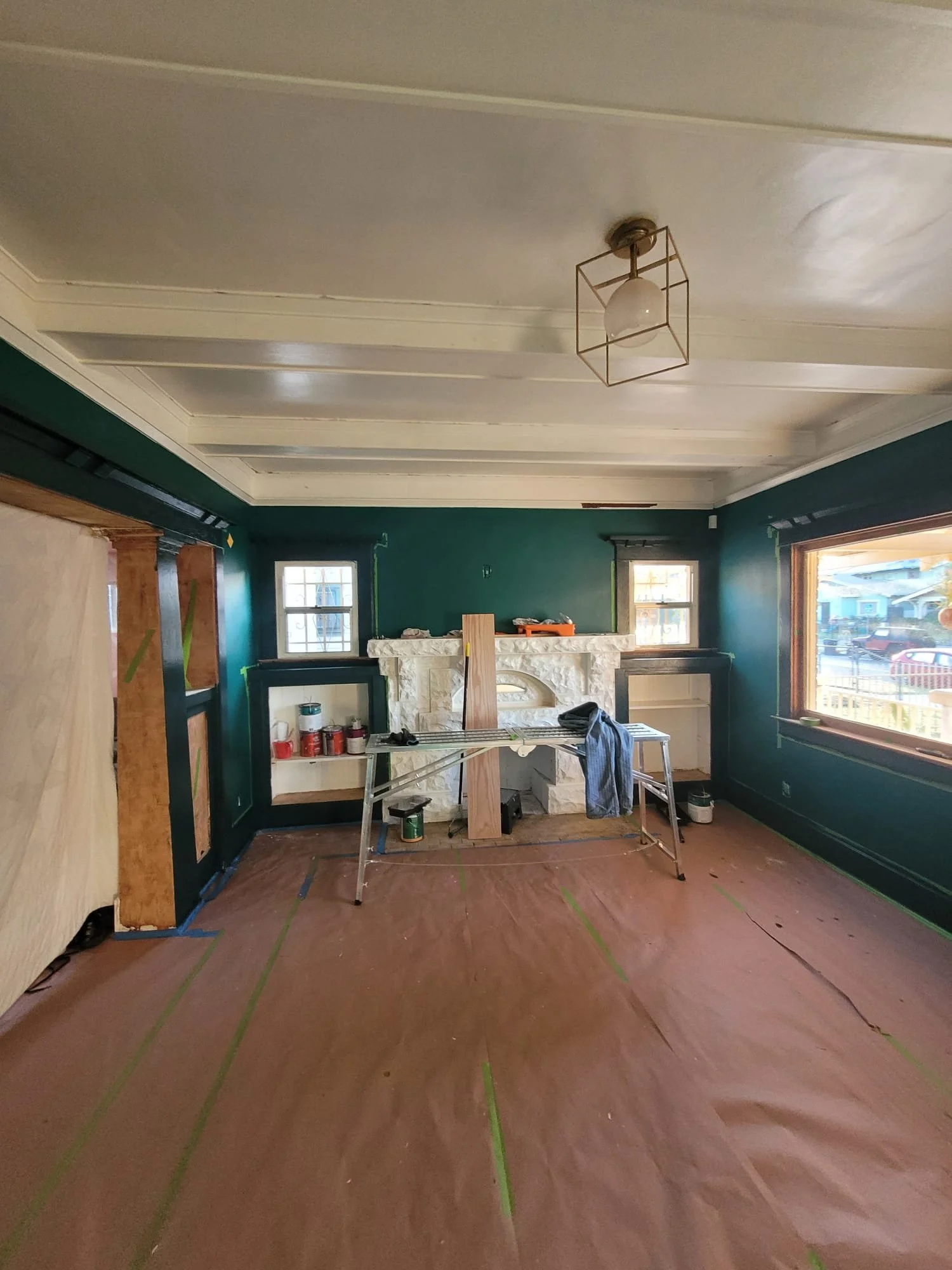 Interior of a room under renovation with a large fireplace, windows, and a ceiling light fixture, with construction supplies and tools scattered around.