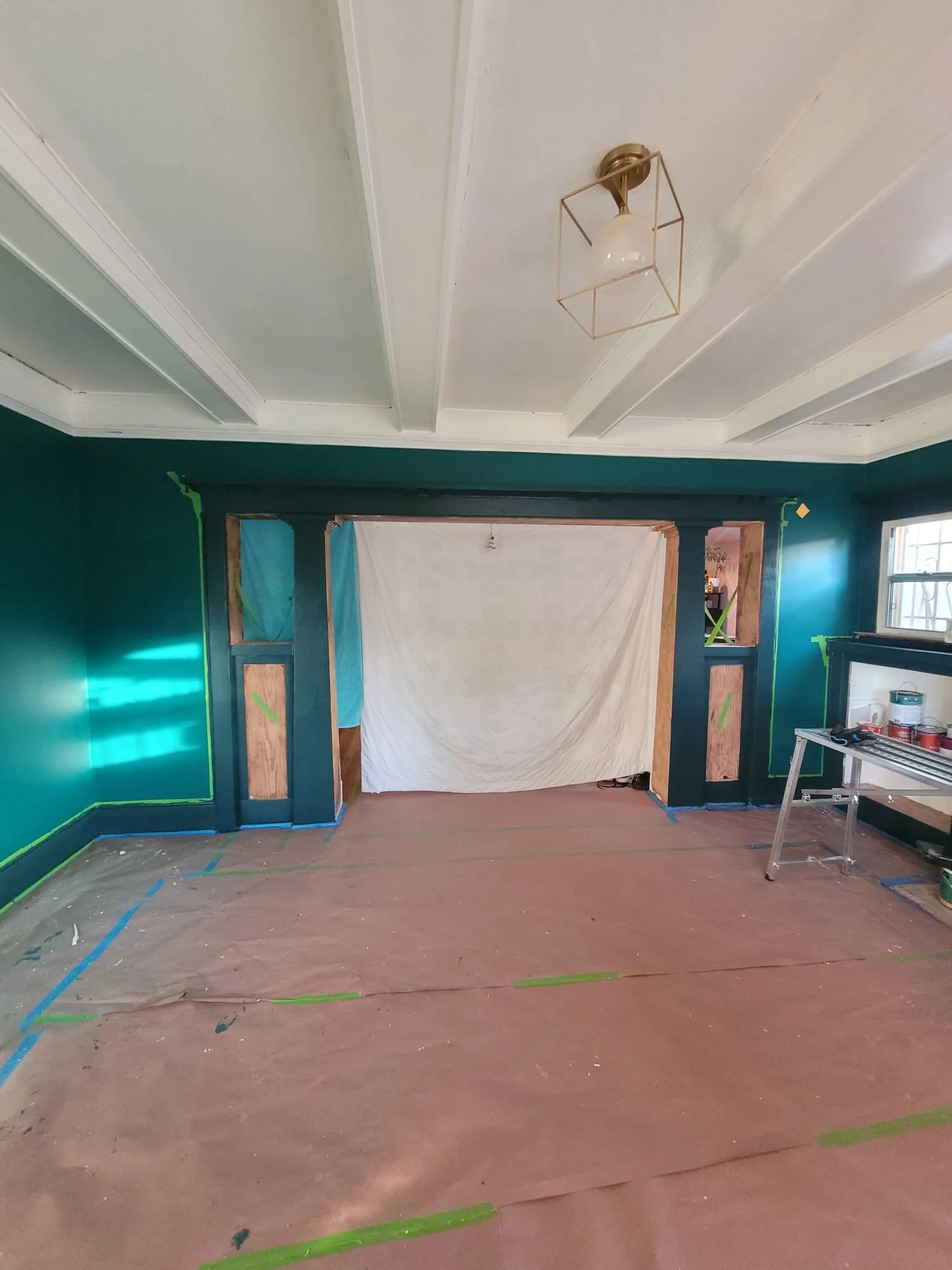 Living room under renovation with green walls, a partially painted ceiling, and a large opening framed in dark blue. Plastic sheeting covers the opening. The floor is covered with brown paper and painter's tape, with painting supplies and a step ladd
