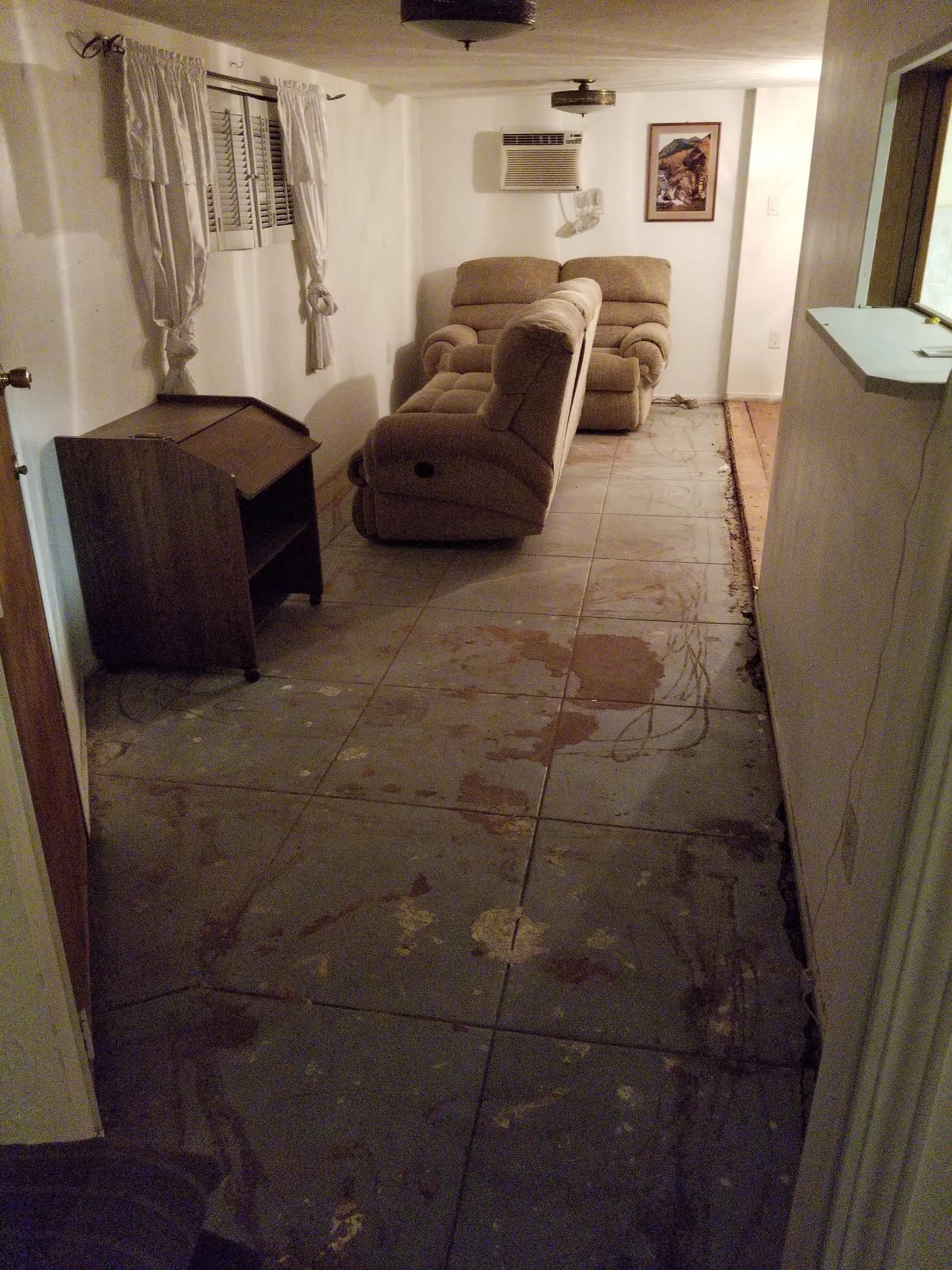 Photo of a living room with beige sofas, a white wall, a window with curtains, and an air conditioning unit. The floor appears to be damaged with visible stains and dirt.