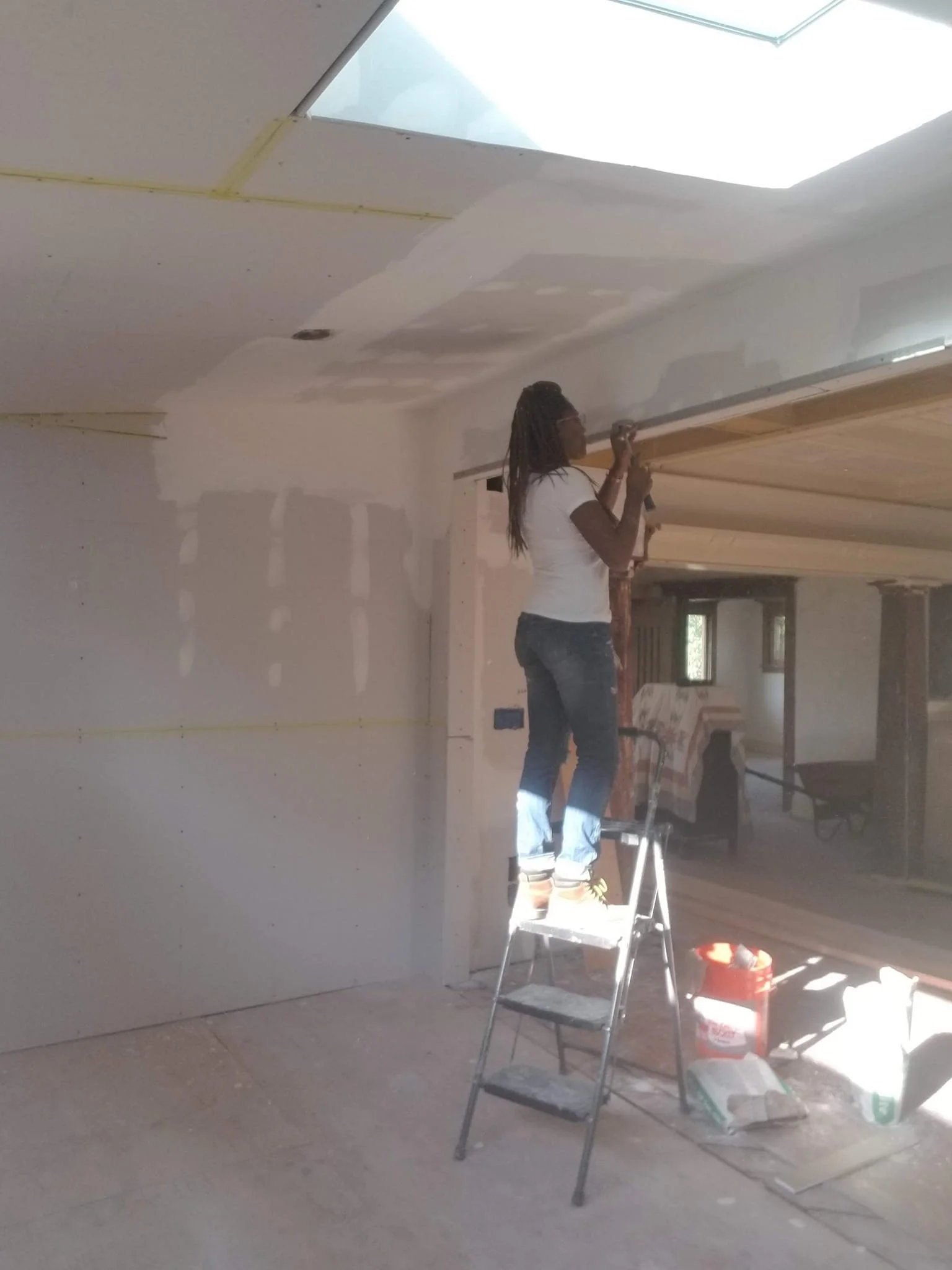 A woman with long hair and glasses is standing on scaffolding, painting or drywalling a ceiling inside a room under construction, with construction tools and supplies on the floor.