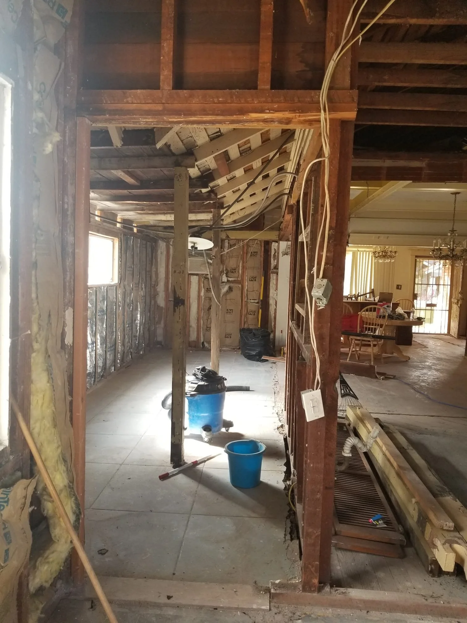 Kitchen undergoing renovation with exposed wooden framing, insulation, and construction materials, including a blue bucket, vacuum, and various tools.