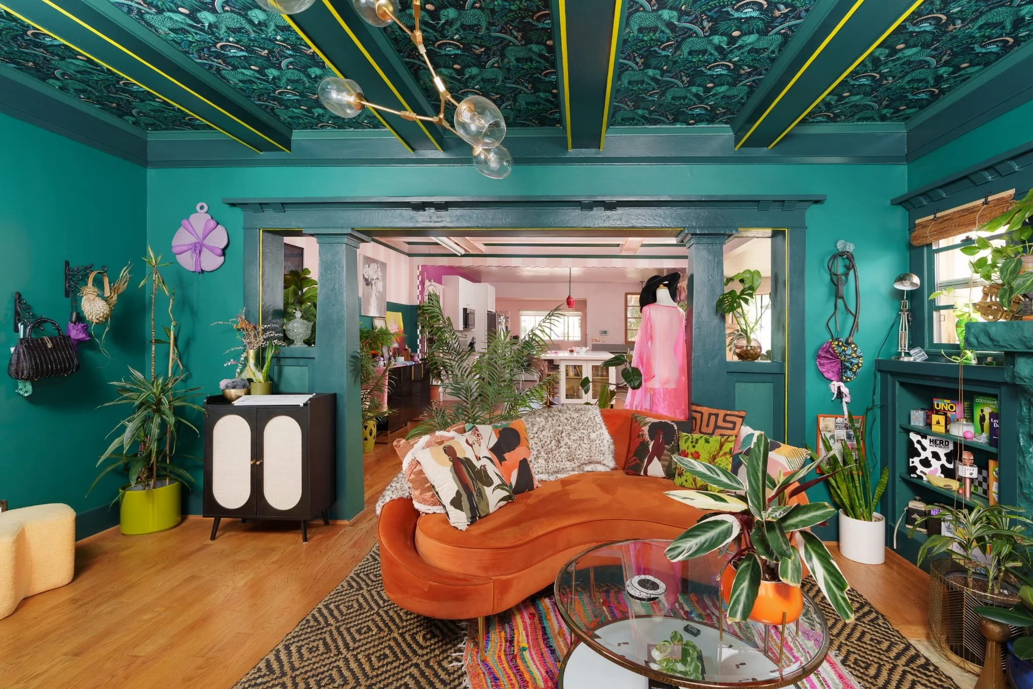 A colorful living room with teal walls and ceiling decorated with a tropical leaf pattern. The space features various plants, a patterned rug, a curved orange sofa with multiple pillows, a glass round coffee table with a decorative plant, and eclecti