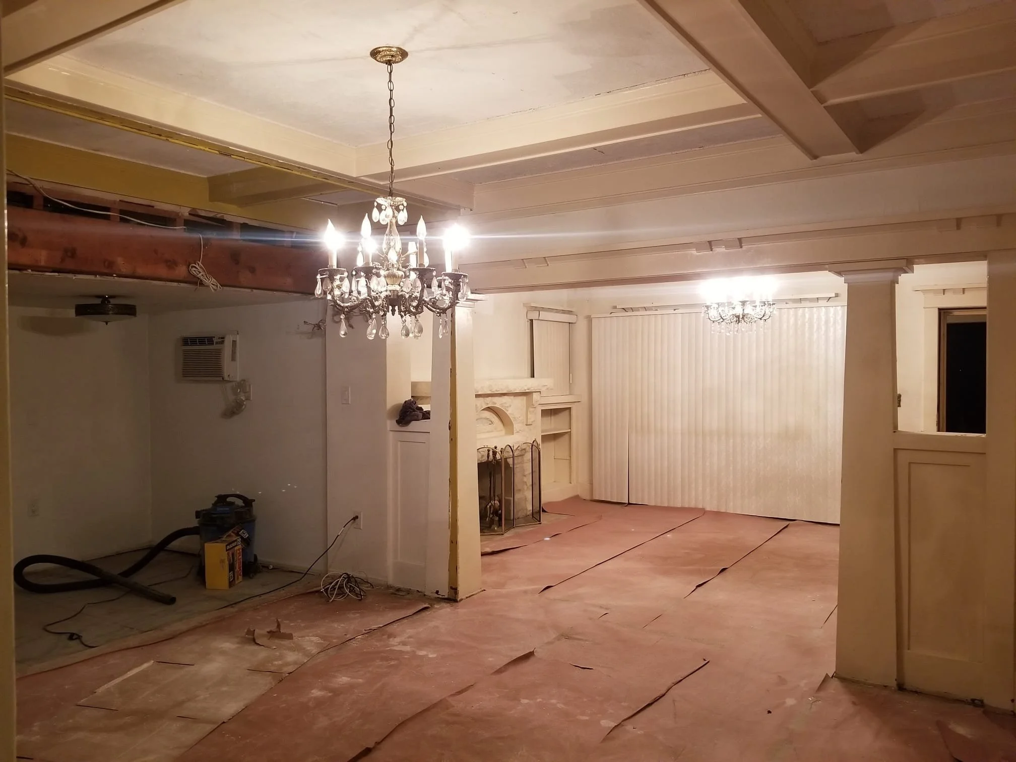 Living room under renovation with unfinished flooring, two chandeliers, and construction tools and materials on the floor.