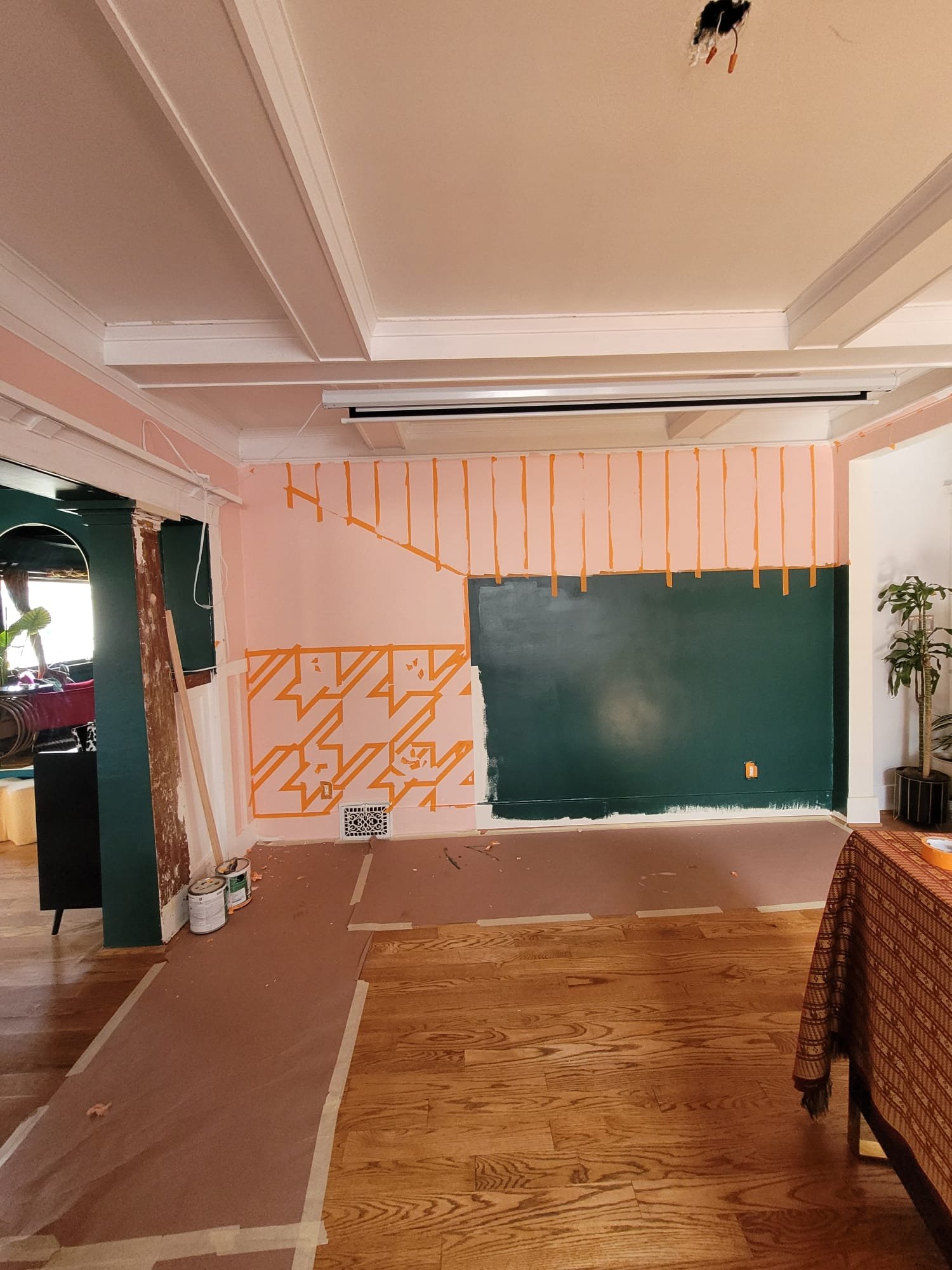 Interior room under renovation with partially painted walls, painting supplies on the floor, drop cloths covering the hardwood floor, and a piece of furniture on the right side. No ceiling light fixtures installed yet.