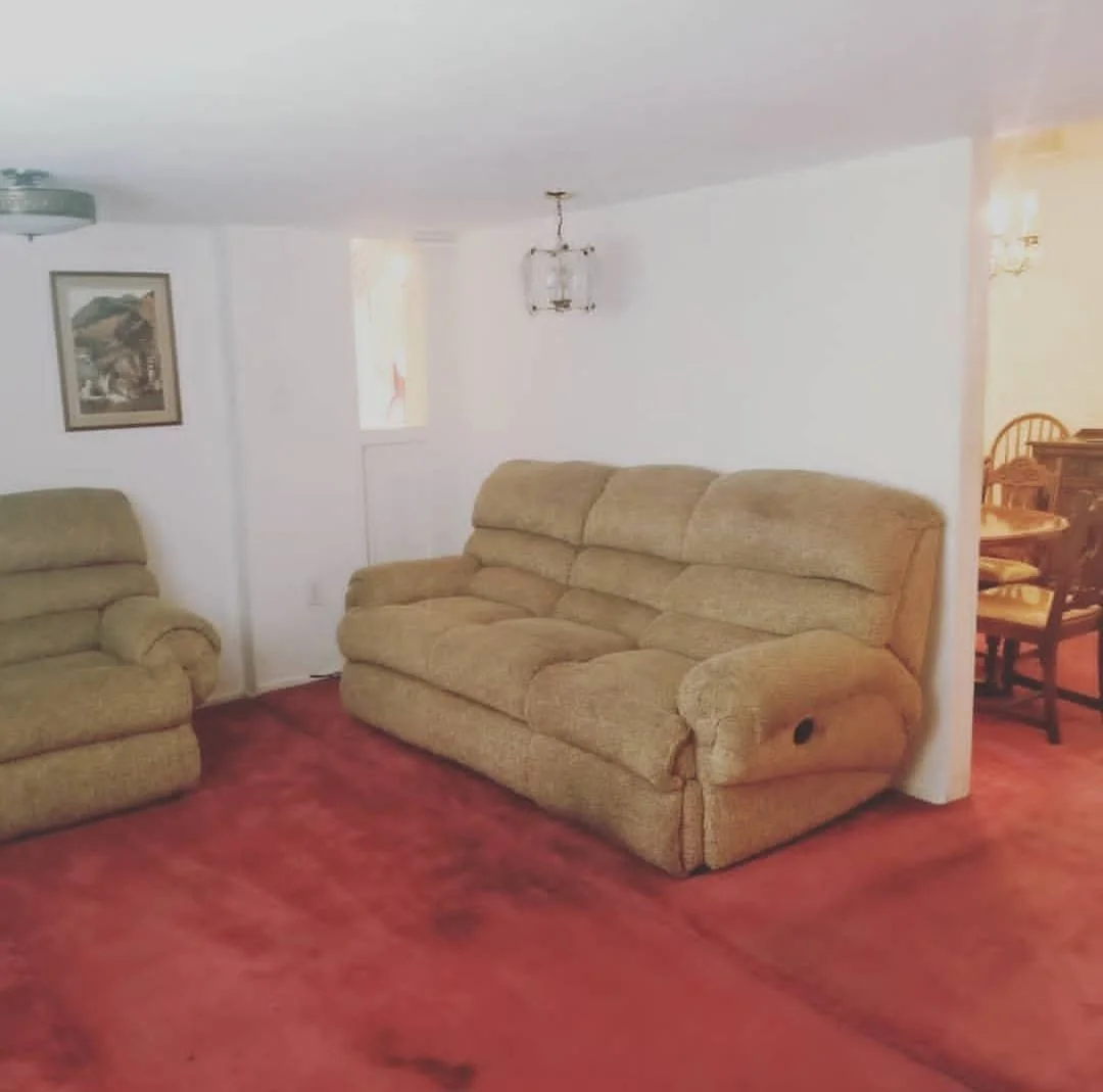 Living room with beige upholstered sofas, red carpeted floor, white walls, framed picture on the wall, small window, and a chandelier hanging from the ceiling. A dining area with wooden table and chairs is visible in the background.