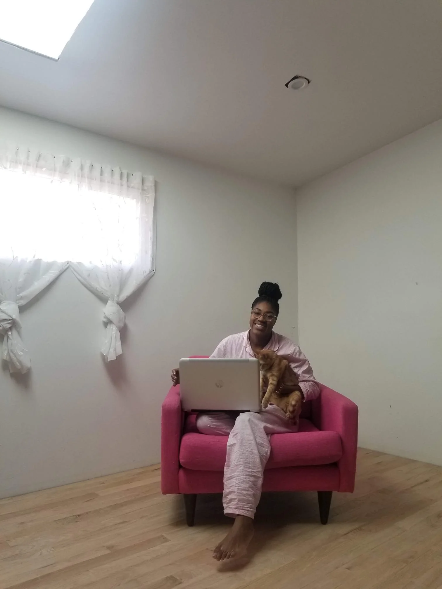 A woman sitting on a pink chair in a room with wooden flooring and white walls, holding a dog on her lap. She is smiling and working on a laptop.