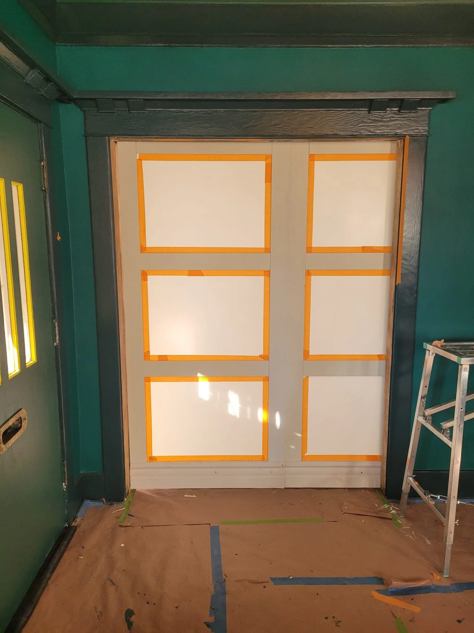 Inside a room with green walls, a door with white panels outlined in orange painter's tape, and a stepladder on the right side. The floor is covered with cardboard and painter's tape.