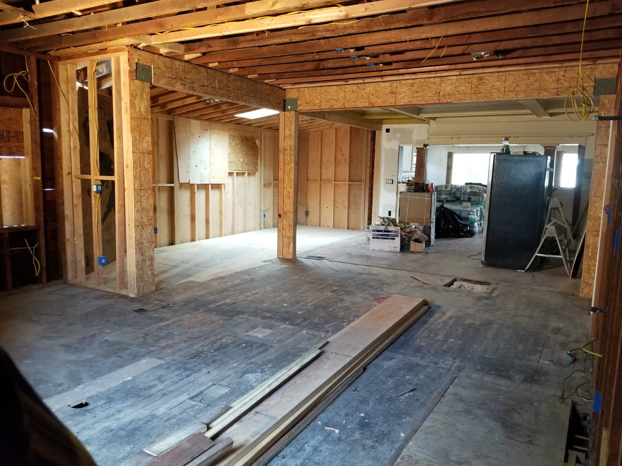 Interior of a house under renovation with exposed wooden beams and framing, construction materials, and tools visible, including wires hanging from the ceiling.