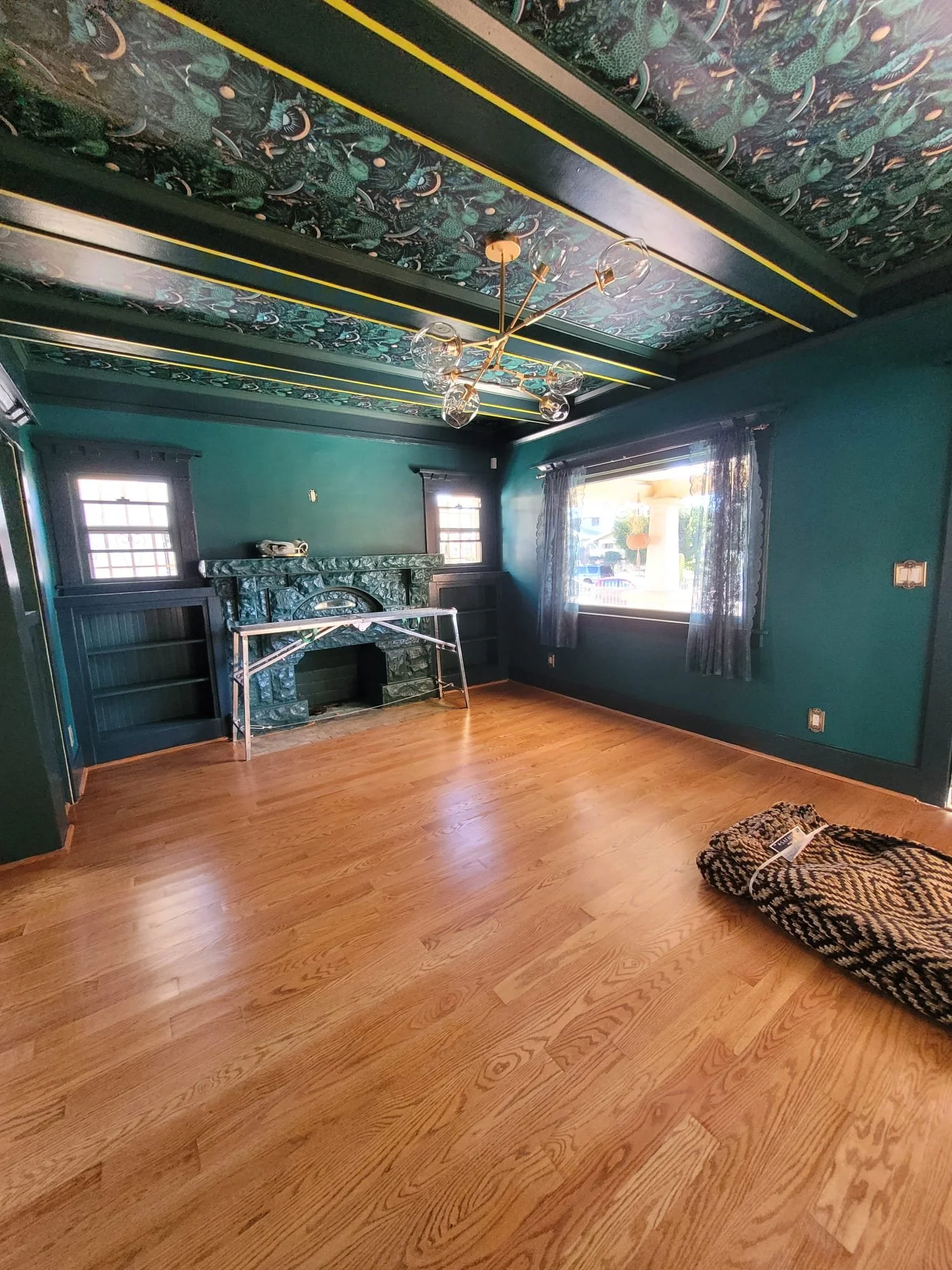 A room with hardwood floors, dark teal walls, and two windows with curtains. There's a green marble fireplace, some fireplace tools, and a pair of slippers on the floor.