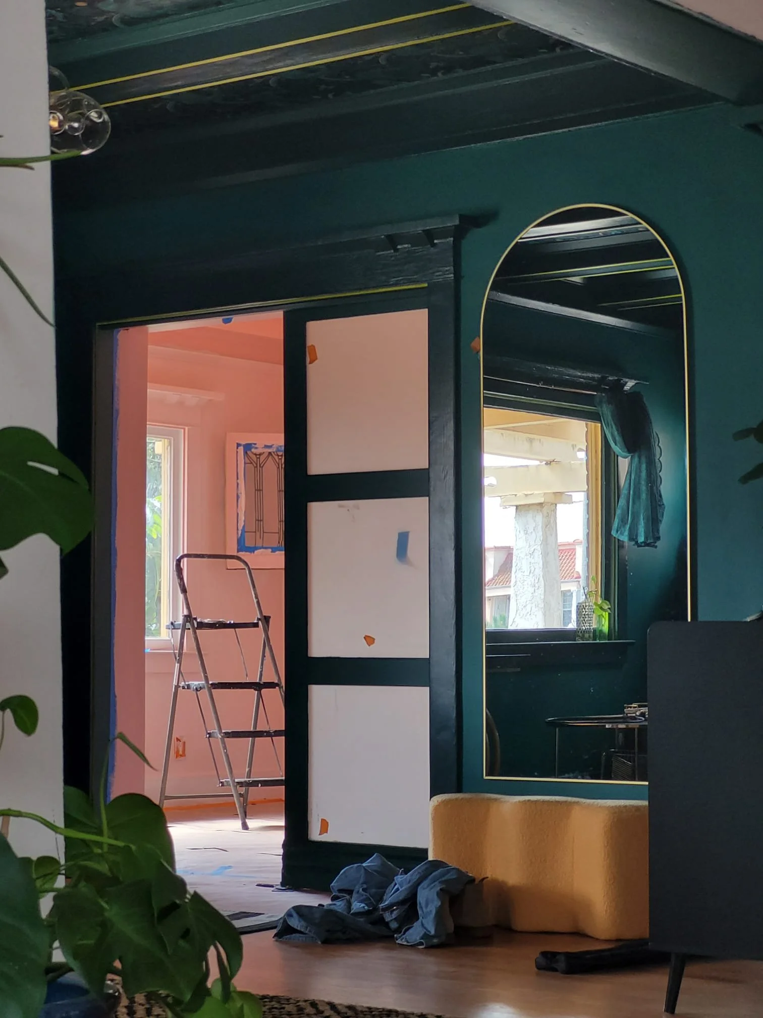 A room undergoing renovation with an open door revealing another room behind it. Inside the room, there is a ladder, a window, and some clothing or fabric on the floor. The walls are painted in warm colors, and there are various objects and furniture
