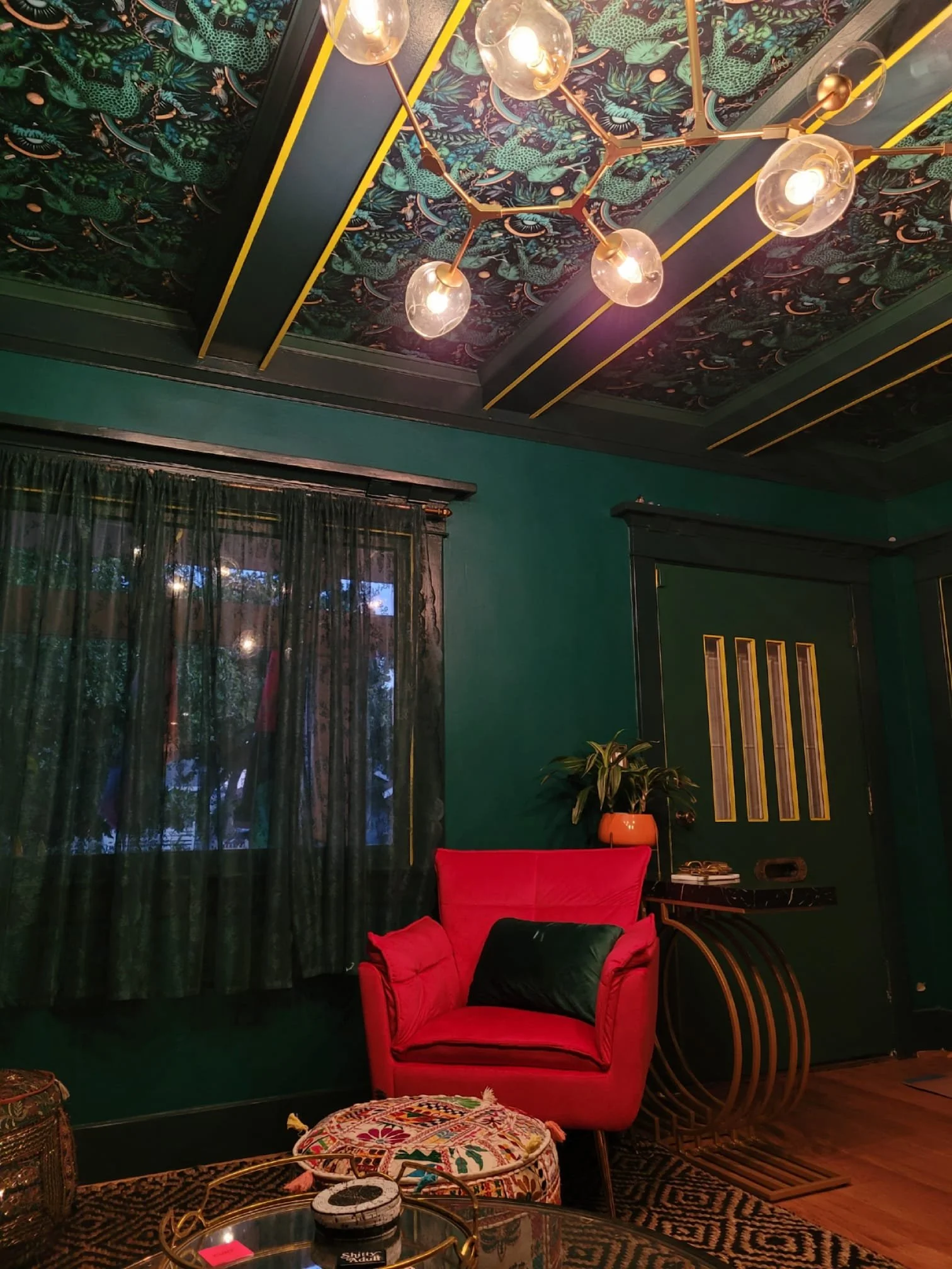 A cozy living room with dark green walls and a window with black curtains. The ceiling has a colorful, tropical-themed wallpaper with pink, green, and blue patterns, and a modern chandelier with bulbs in clear glass globes. There is a bright red armc