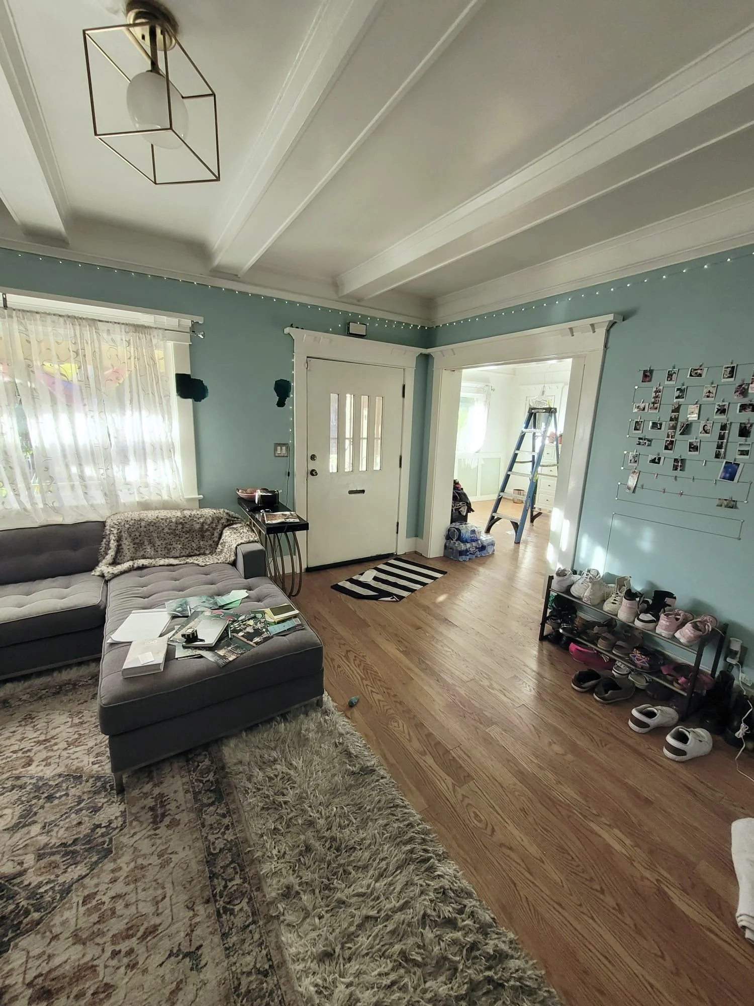 Living room with teal walls, white trim, a window with floral curtains, a gray sectional sofa with a blanket, a shag rug, a small table with magazines, and a shoe rack with shoes. There is a doorway to another room with a ladder and water bottles.