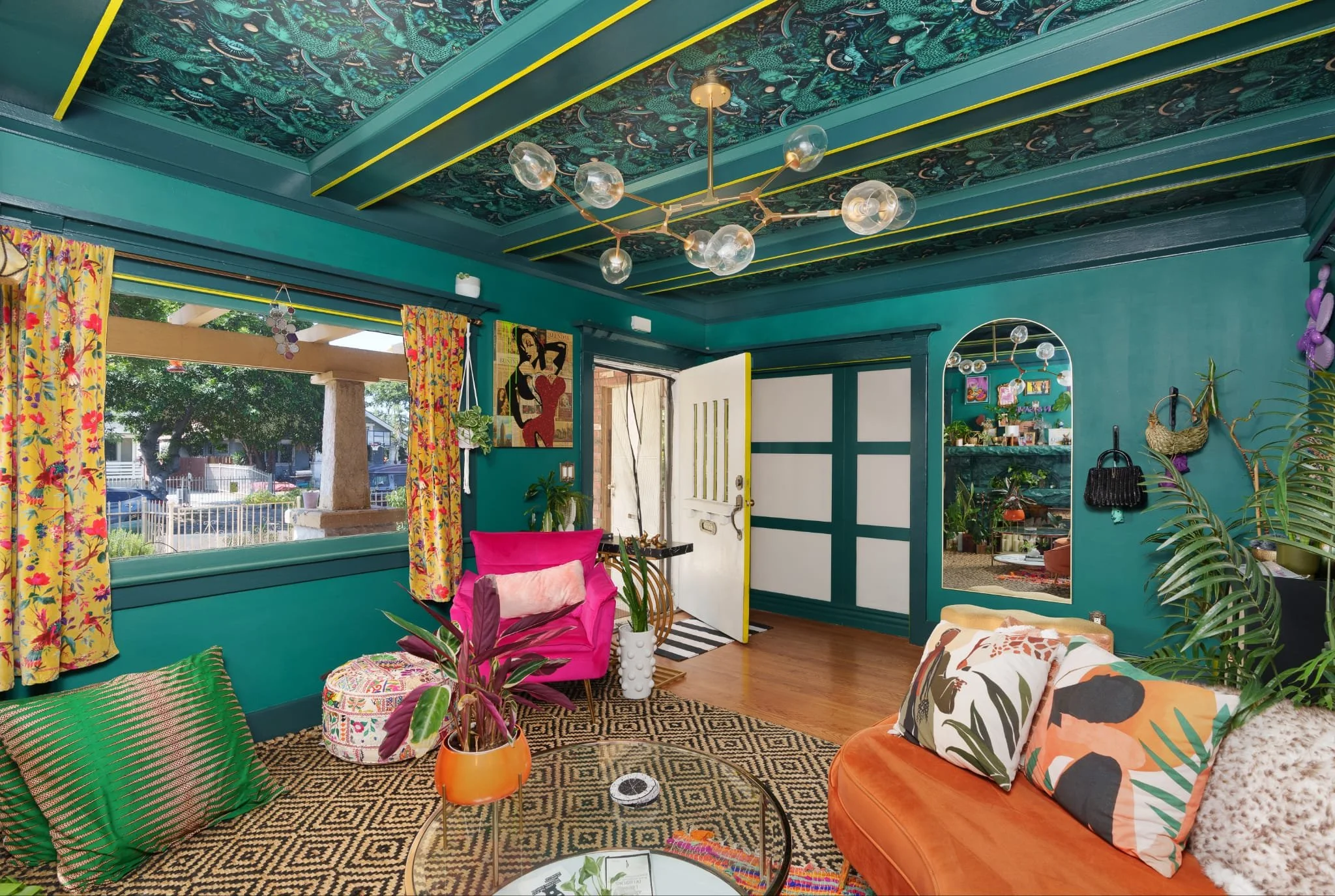 A vibrant living room with teal walls, colorful floral curtains, and eclectic decor, including plants, unique pillows, and modern lighting fixtures.