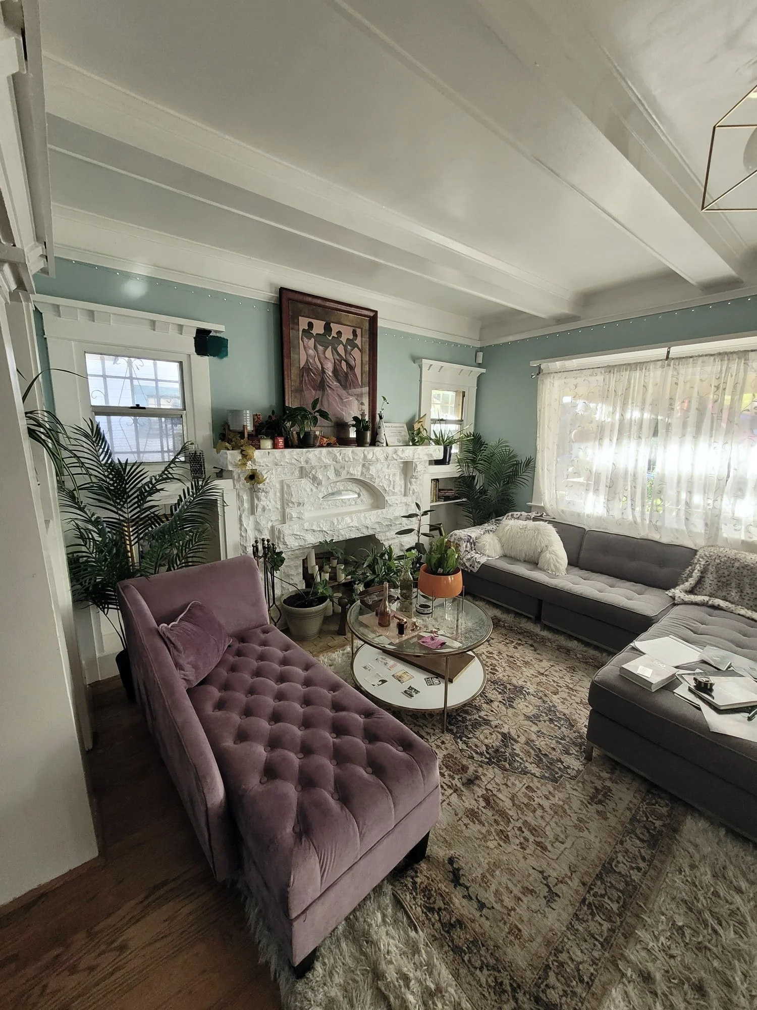 A living room with a pink tufted velvet sofa, a gray sectional sofa, and a round glass coffee table on a patterned rug. There are several potted plants, a fireplace with decorative items, a framed artwork, and large windows with sheer curtains.