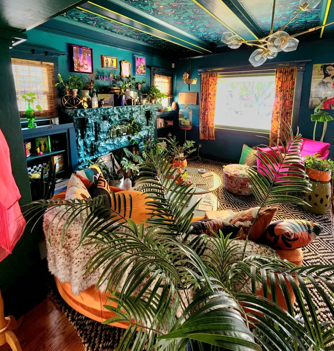 A colorful living room with teal walls, a patterned ceiling, and large windows with floral curtains. The room is decorated with various plants, vibrant furniture, framed artwork, and eclectic decor including a teal marble fireplace and a plush orange