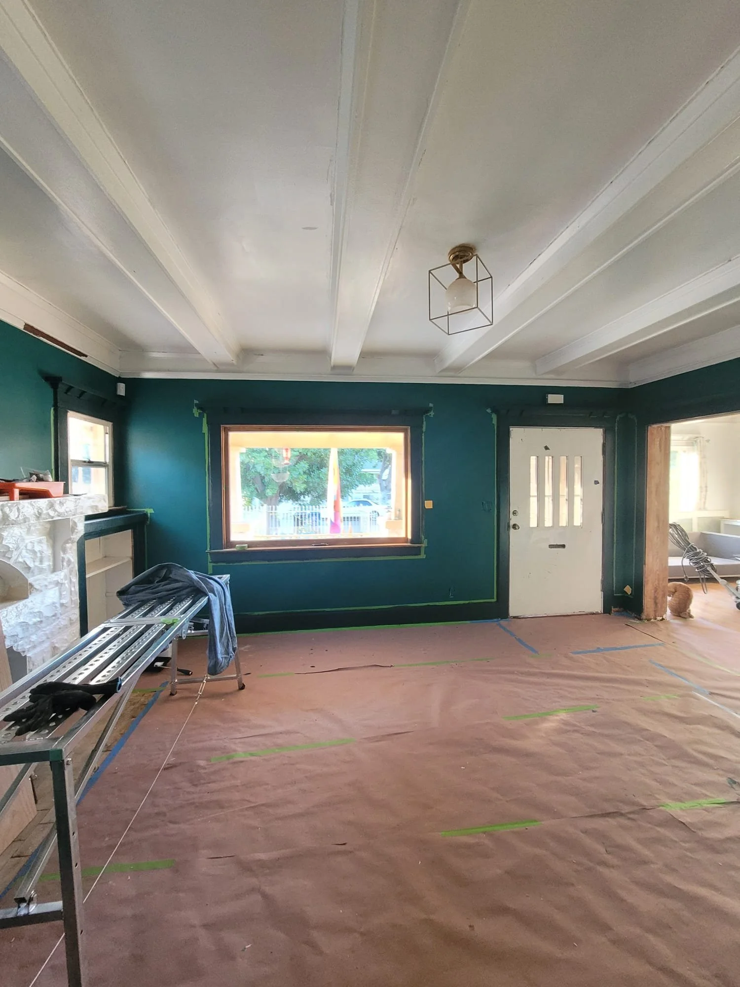 Interior of a room under renovation with a green wall, a window, and construction tools on a table. The floor is covered with brown protective paper, and the ceiling has decorative beams with a central light fixture.