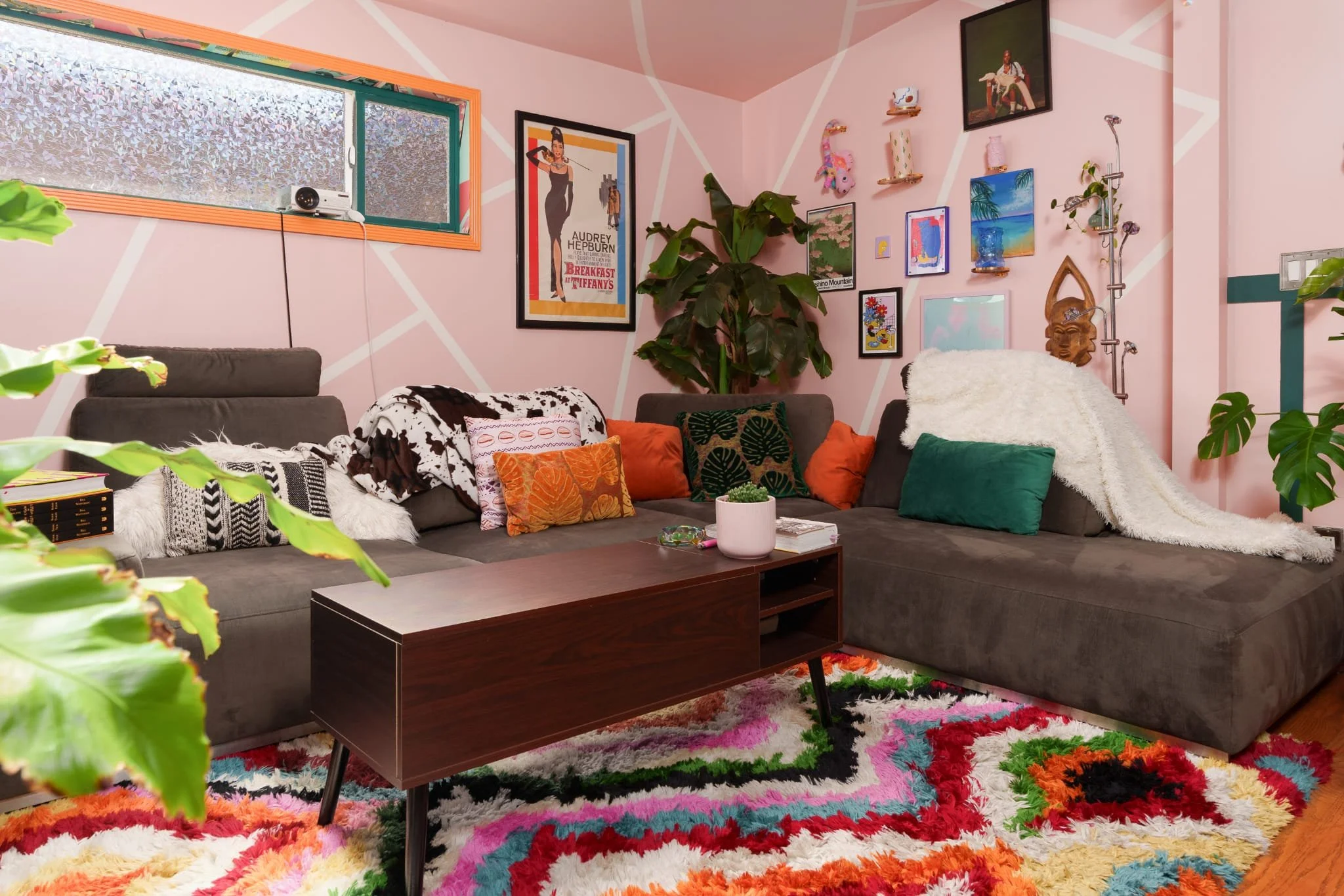 Living room with pink walls decorated with eclectic artwork, plants, and a multicolored rug. Gray sectional sofa with orange and patterned throw pillows, a wooden coffee table, and a clothing draped over the side.