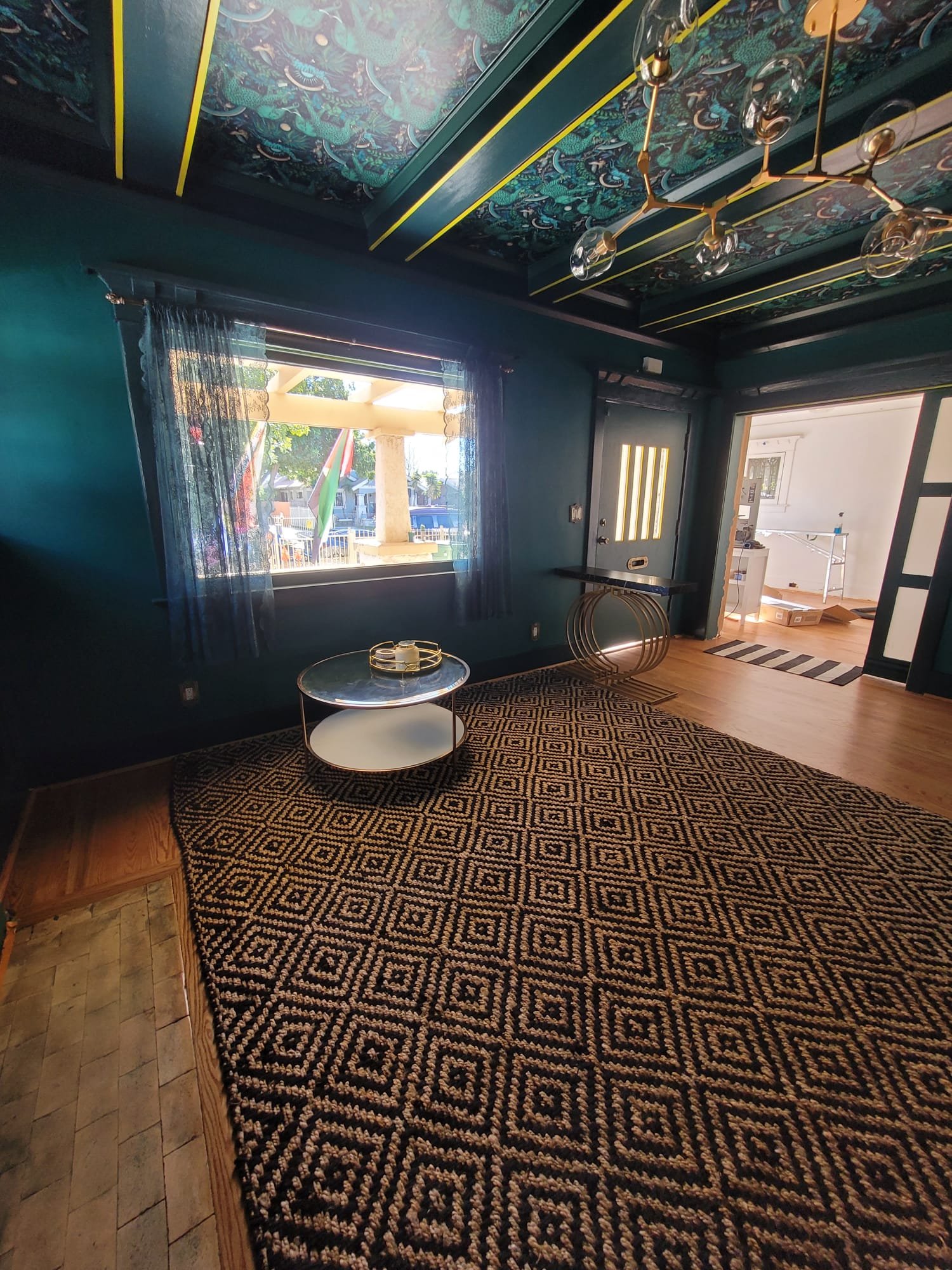 Interior of a room with dark teal walls and a patterned carpet. There is a window with sheer curtains and a view of a porch outside. A geometric chandelier is hanging from the decorated ceiling, and a small round table with decorative items on it is 
