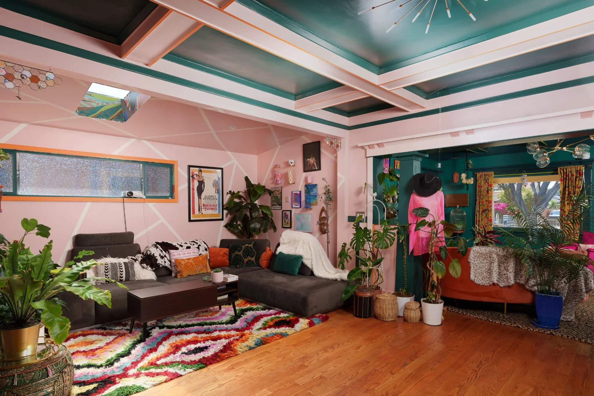 A cozy, colorful living room with pink and teal walls, decorated with various artwork and houseplants. There is a large dark gray sectional sofa with patterned and solid cushions, a wooden coffee table, and a rainbow-colored shag rug on a wooden floo