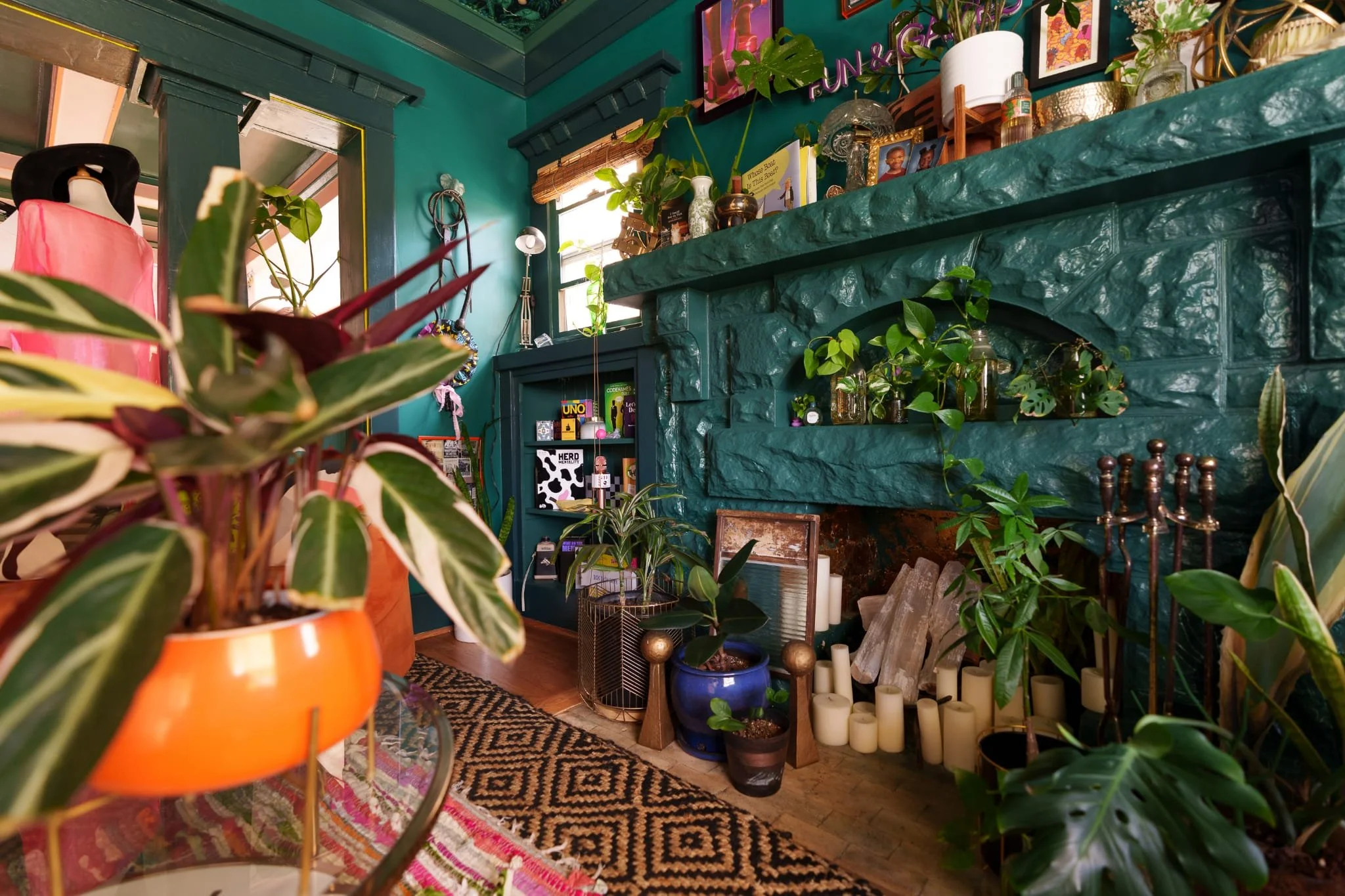 Indoor space decorated with green walls, filled with various potted plants, candles, and decorative items, including framed artwork and books, creating a cozy, eclectic atmosphere.