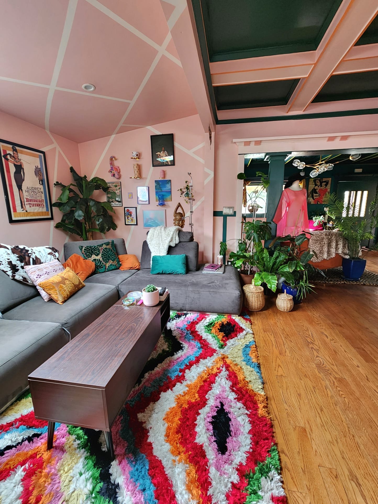 A colorful, shaggy area rug dominates a living room with a gray sectional sofa decorated with various patterned and textured pillows. The wall behind the sofa is decorated with framed posters, photographs, and small shelves holding decorative items. 