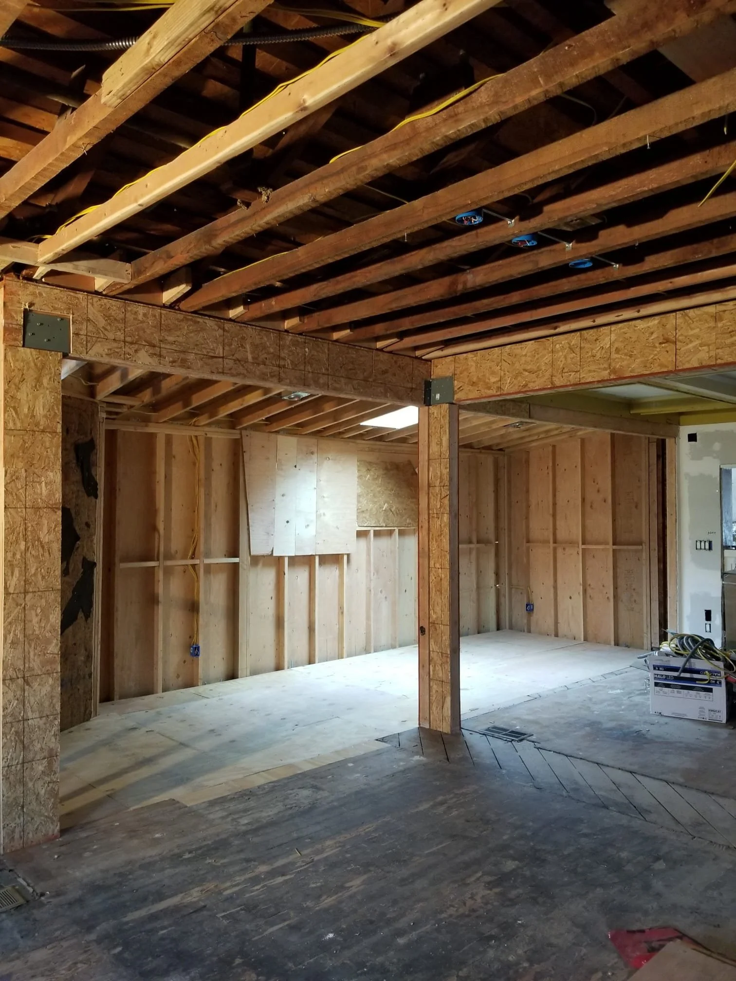 Interior of a room under construction with exposed wooden framing, overhead electrical wiring, and partially installed drywall. There are construction tools and materials on the floor.