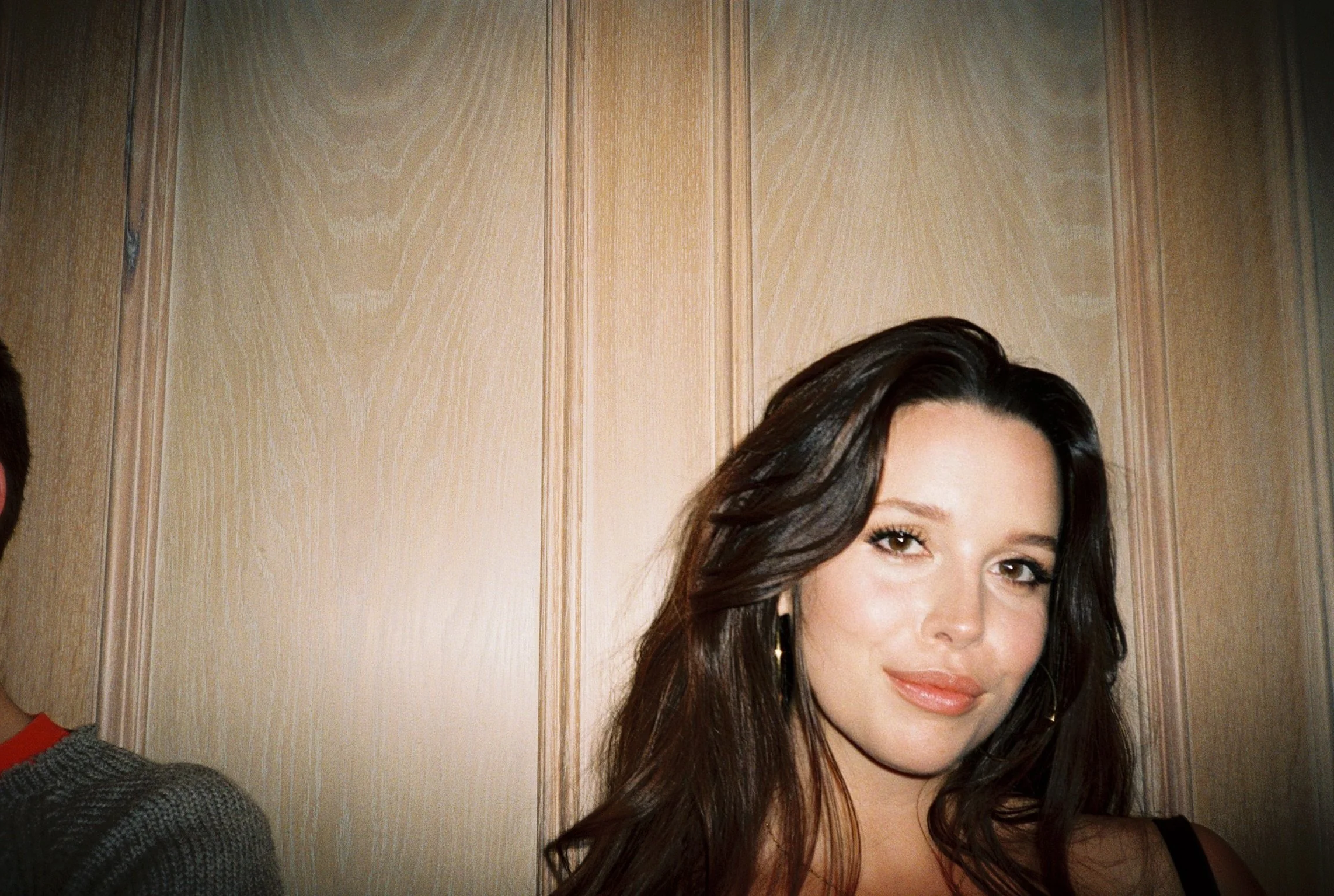 A woman with dark, wavy hair and a slight smile. She is indoors, standing in front of a wooden-paneled wall.