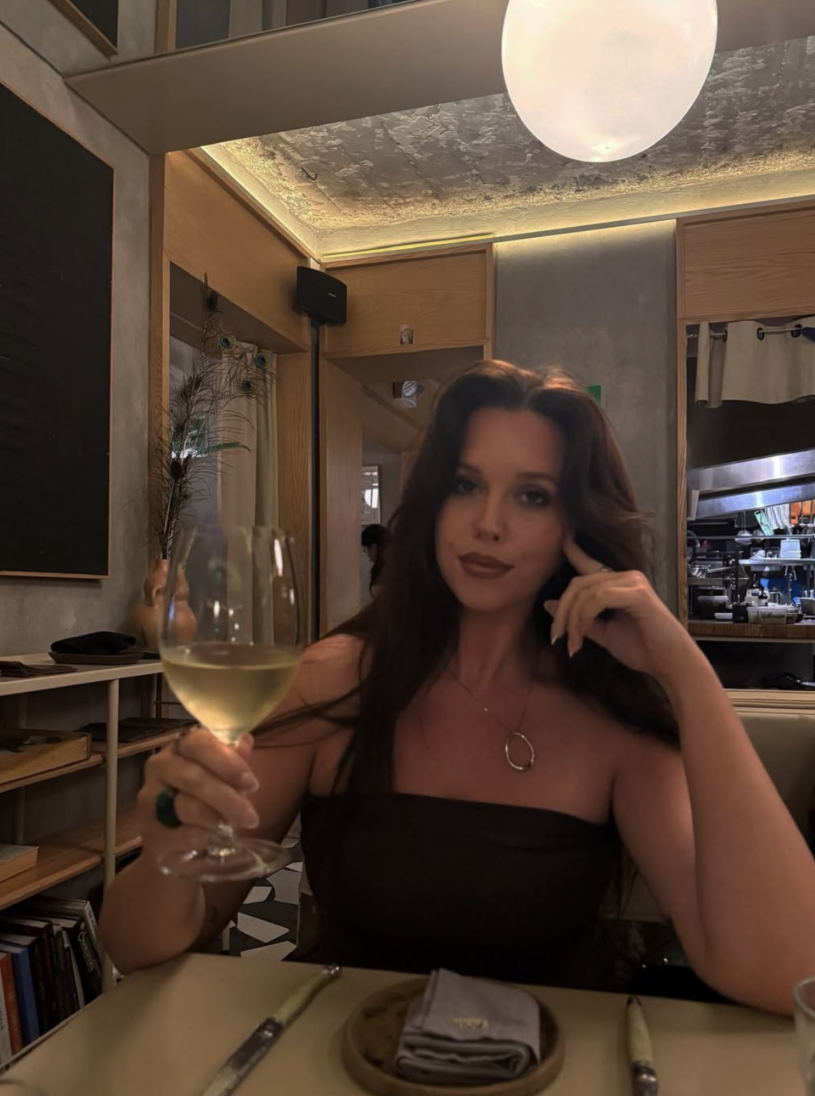 A woman sitting at a restaurant table, holding a glass of white wine, with dark hair, wearing a black strapless top and jewelry, in a modern, warmly lit dining space.