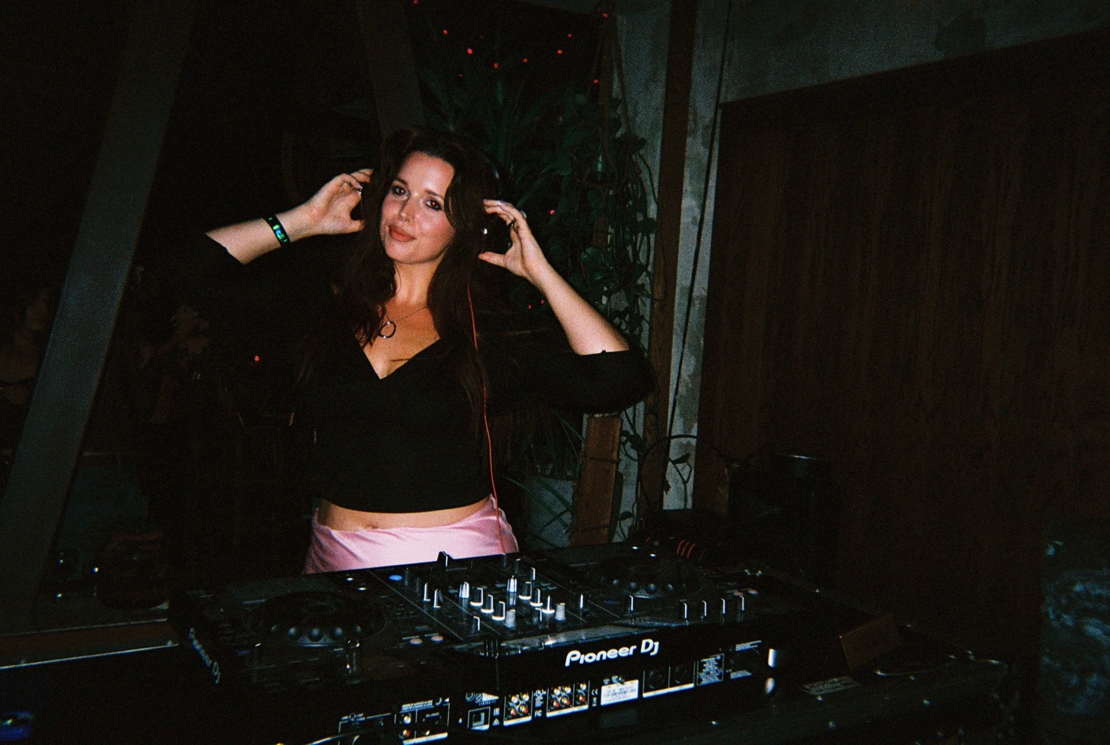 A woman wearing headphones and a black top stands behind DJ equipment, in a dimly lit setting.