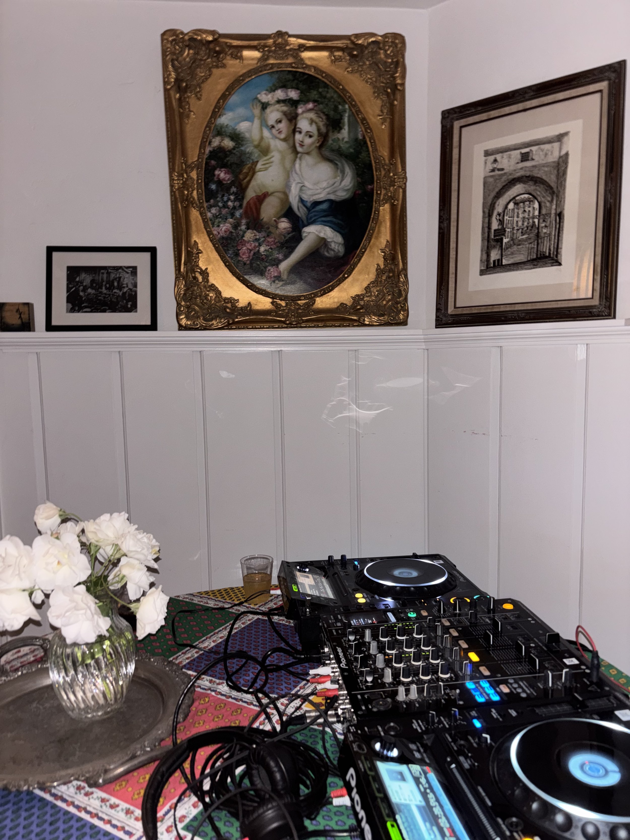A DJ setup with two turntables and a mixer on a colorful tablecloth, with white flowers in a glass vase on the side. There are various wires connected to the equipment, and a small glass of drink next to the setup. In the background, framed artwork hangs on a white wall, including a large classical-style painting of two women with cherubs, and two smaller black and white pictures.