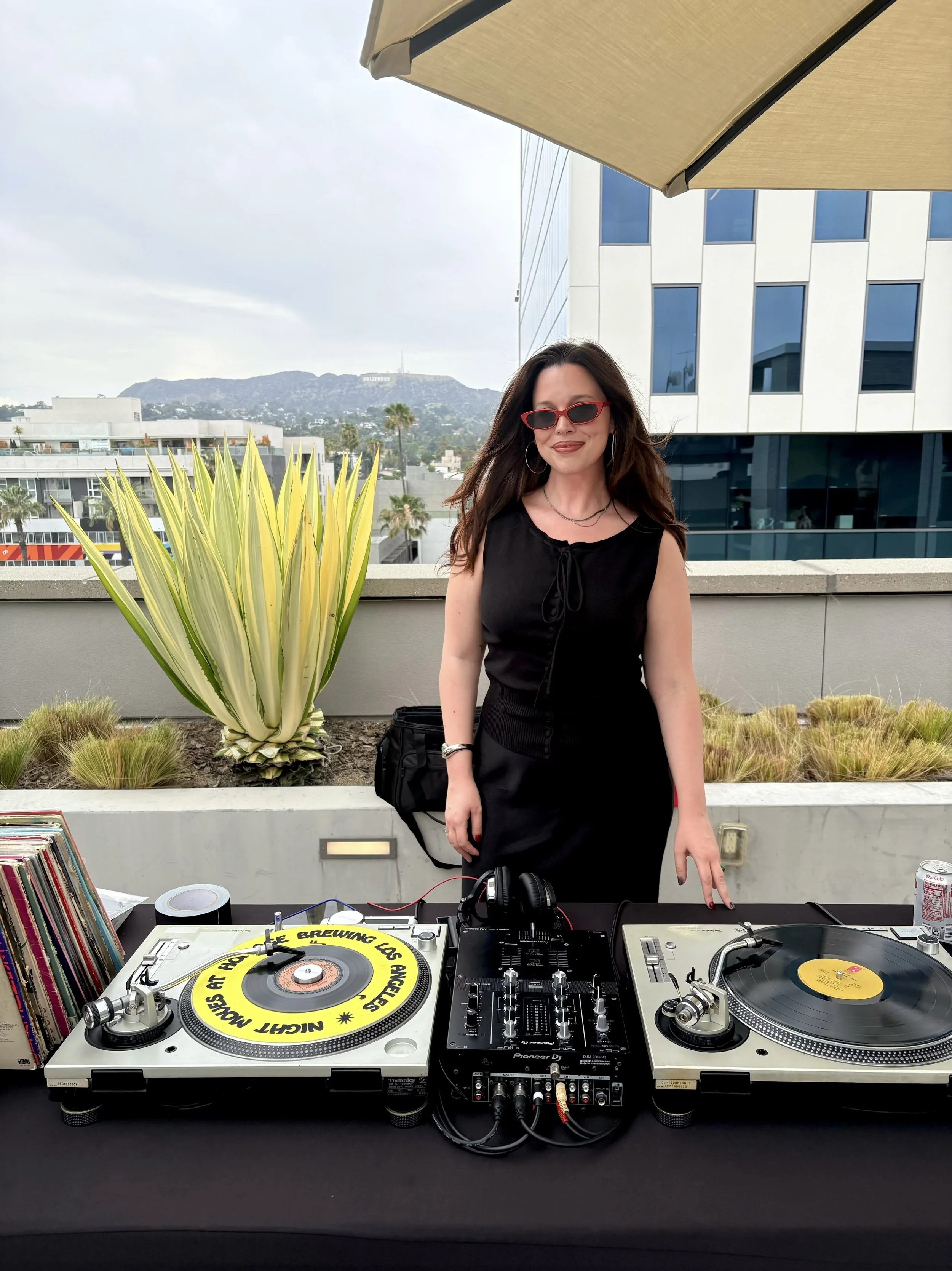 A woman with long dark hair and red sunglasses standing behind DJ turntables and audio equipment on a table with a cityscape and the Hollywood sign in the background.