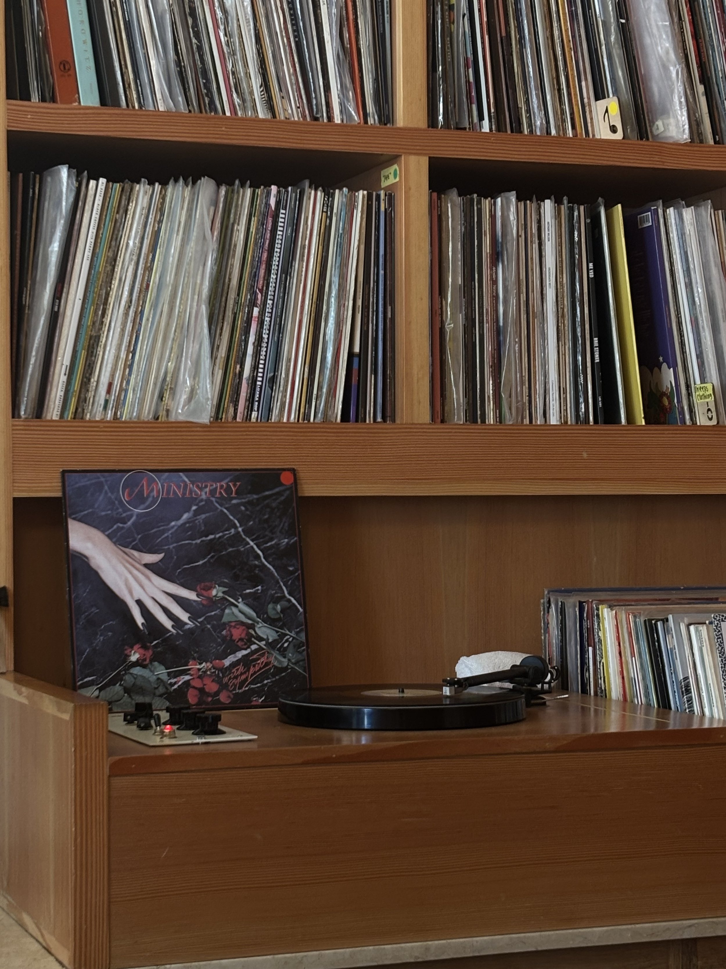 A wooden bookshelf filled with vinyl records, with some records in plastic sleeves, and a turntable on the lower shelf.
